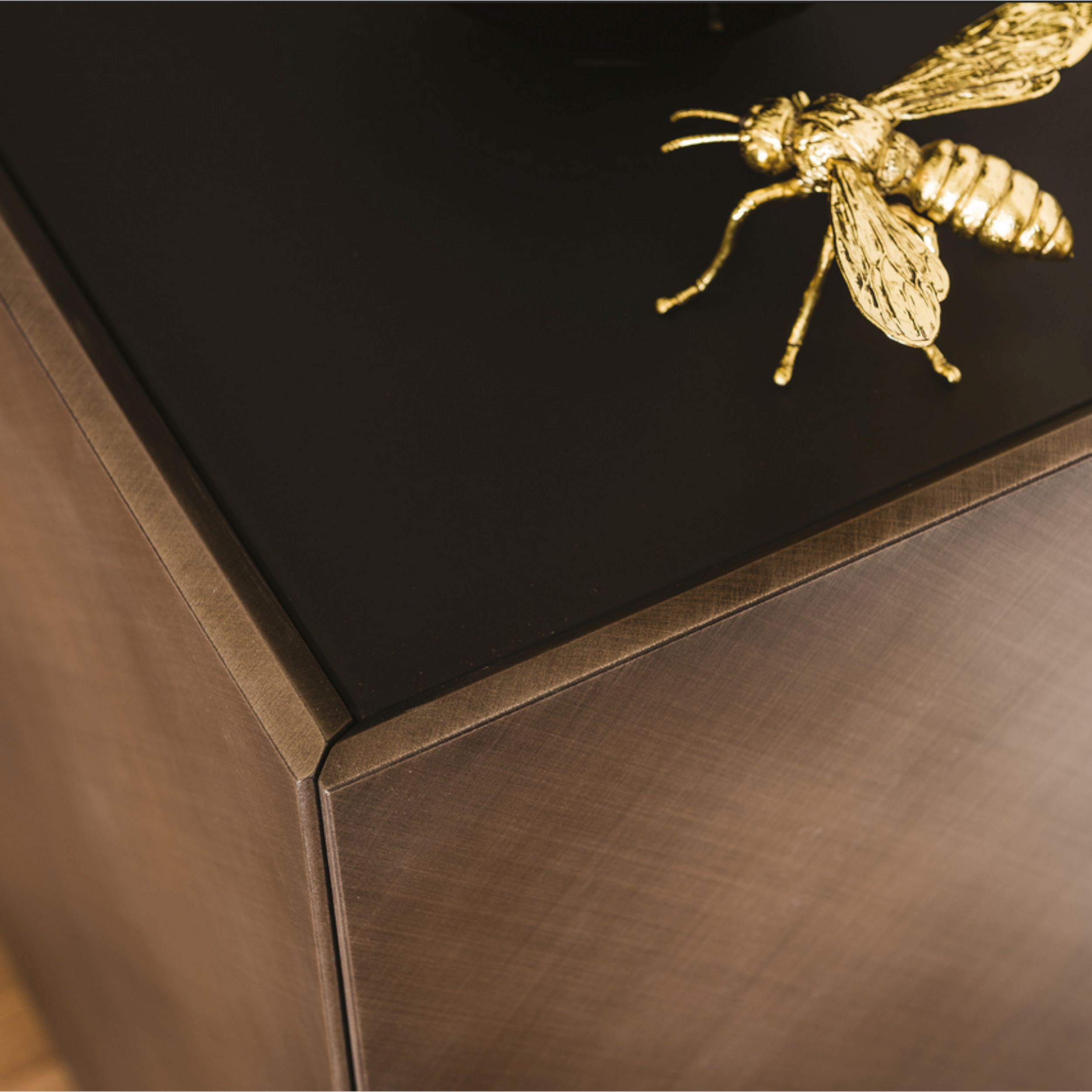 Zu sehen ist in Nahaufnahme von schräg oben die Ecke eines Sideboards mit schwarzer Abdeckplatte und bronzefarbener Front; auf dem Sideboard platziert ist die Figur einer goldenen Biene.