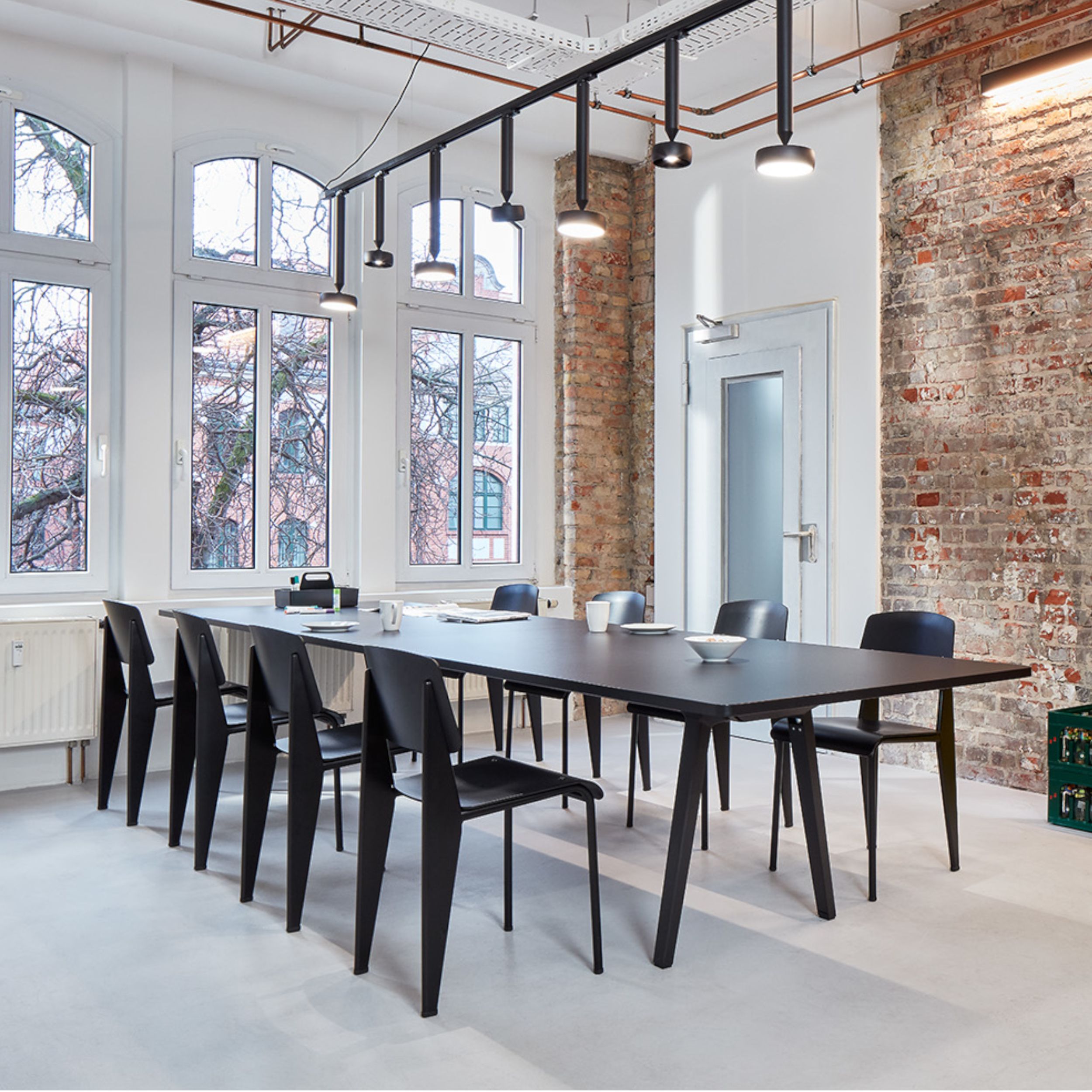 Das Bild zeigt ein modernes Loft mit hoher Decke, Backsteinwand und Rundbogenfenstern. Im Zentrum steht ein schwarzer Konferenztisch mit acht dunklen Standard SP Stühlen von Vitra.