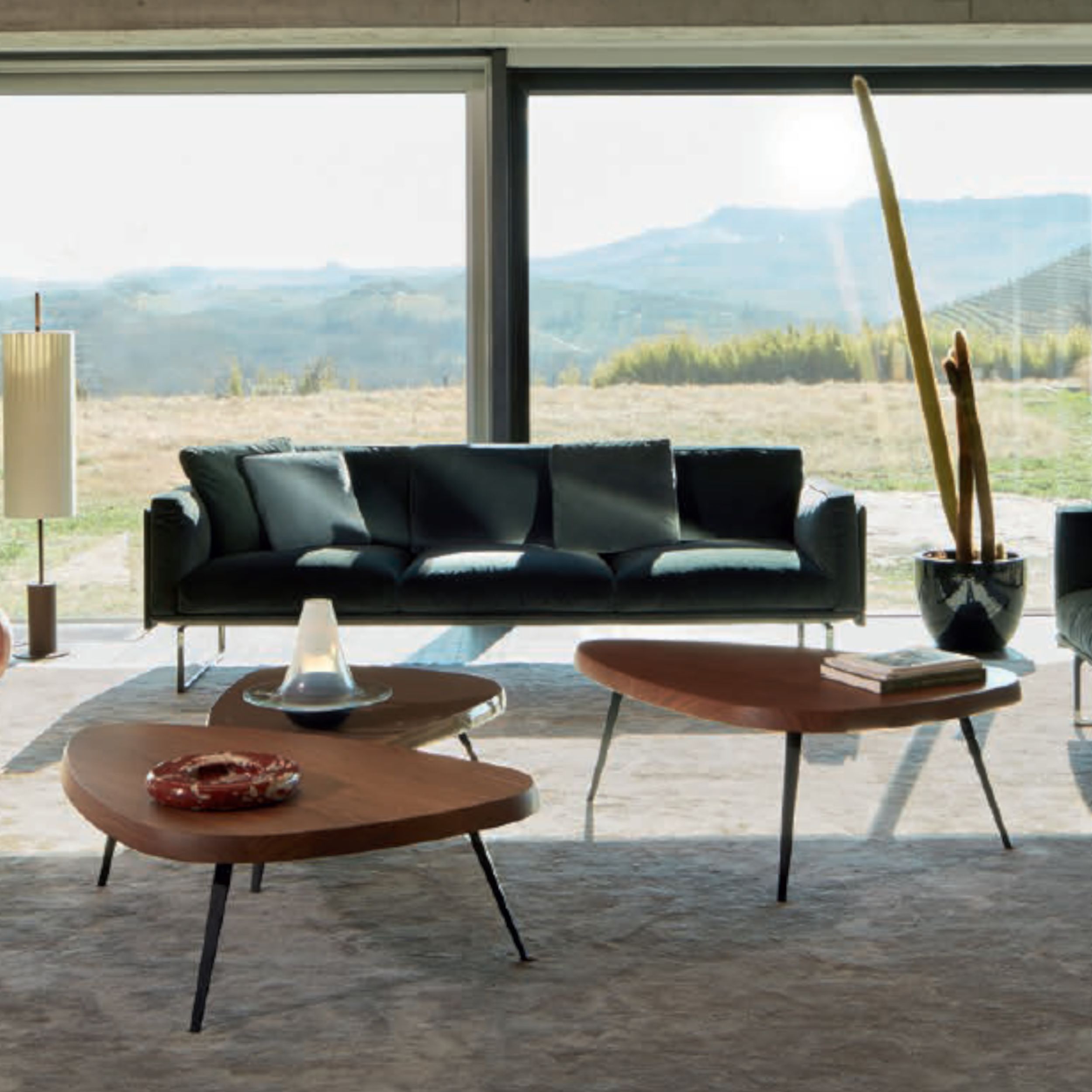 Drei Couchtische "Mexique" von Cassina, kombiniert mit einem blaugrünen Sofa und einem hellen Teppich, stehen in einem großzügigen Wohnzimmer vor einem Panoramafenster.