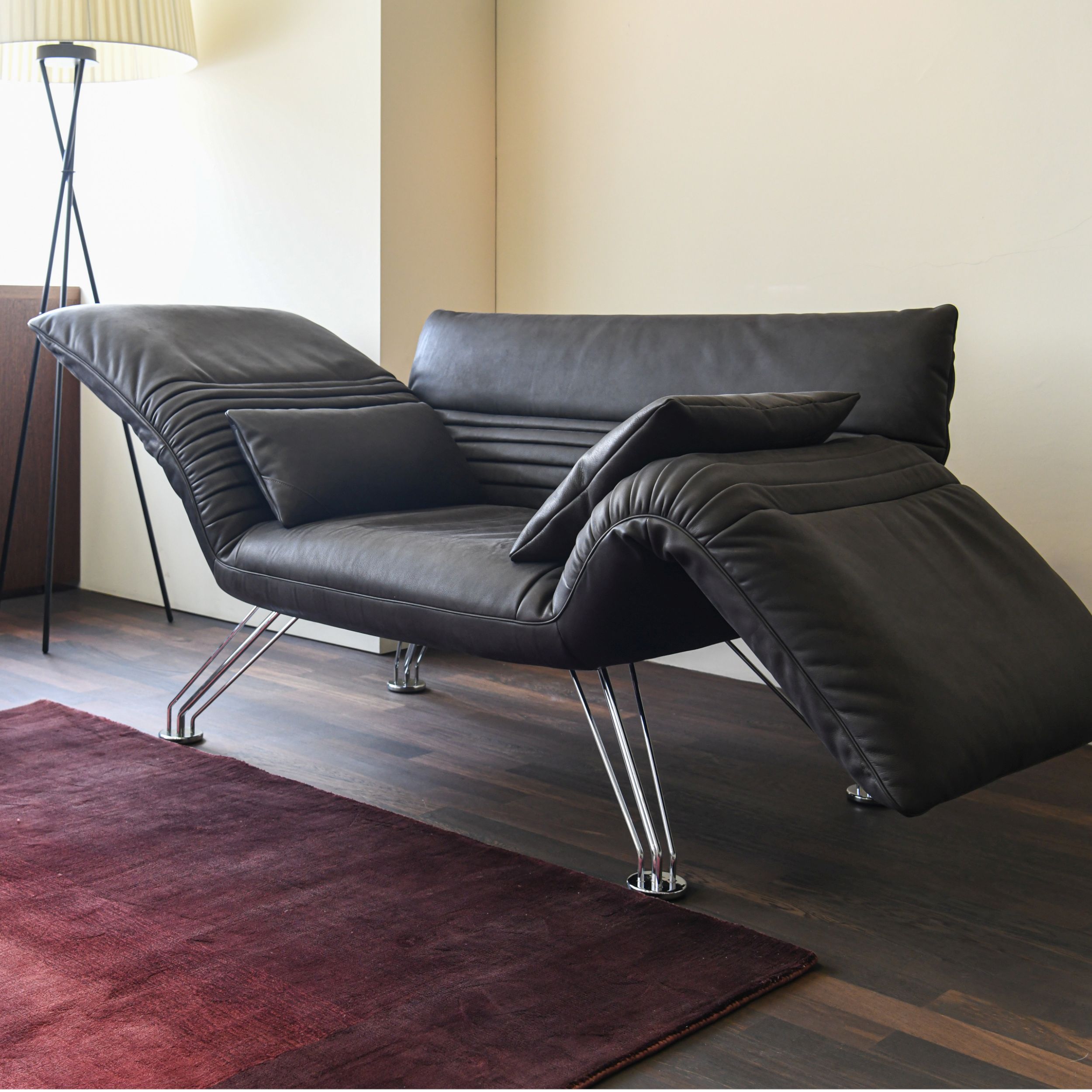 Eine moderne Relaxliege in schwarzem Leder; Mpodell DS- Wing von De Sede, steht auf dunklem Parkettboden, ergänzt durch einen violetten Teppich und eine Stehleuchte.
