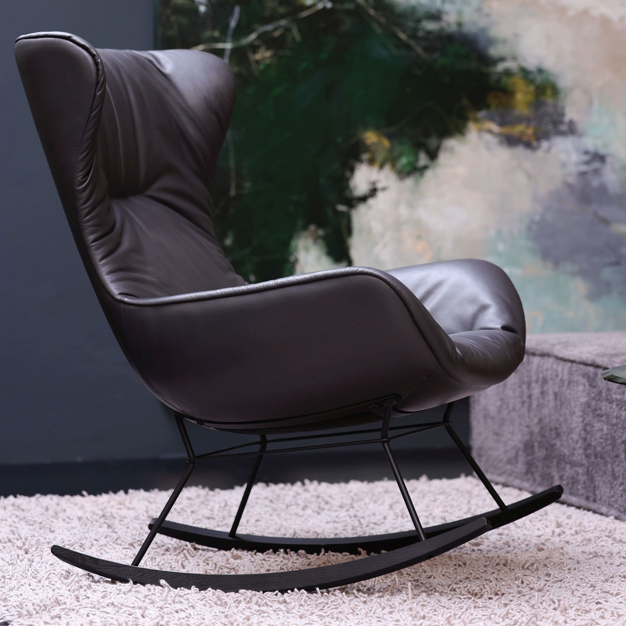 Schaukelstuhl Leya Rocking Wingback Chair in anthrazitgrauem Leder in seitlicher Ansicht in einem elegenten Interieur.