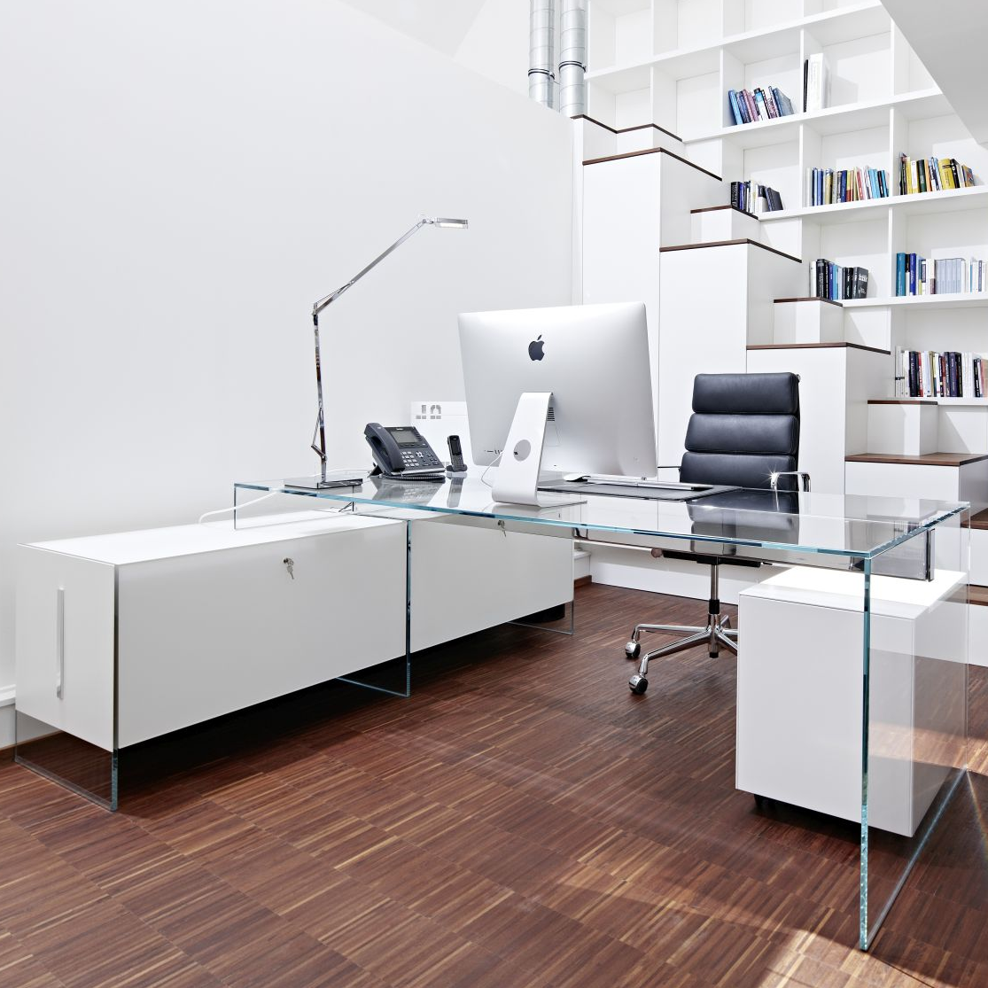 Büro mit Glasschreibtisch, Bürostuhl und Rollcontainer vor einem Bücherregal