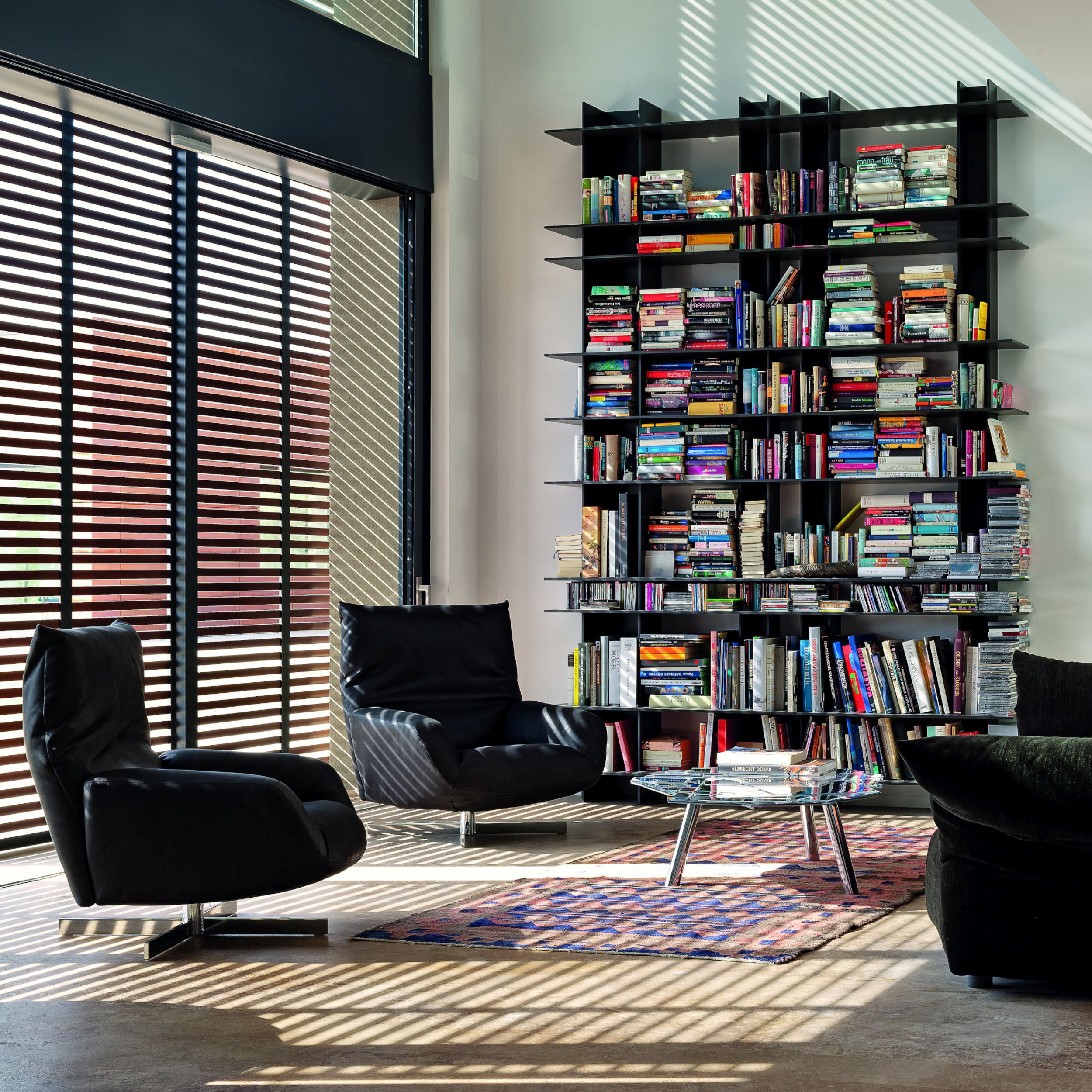 Das Bild zeigt ein Wohn- oder Lesezimmer mit hoher Decke und einem großen, schwarzen Bücherregal. Links im Bild stehen zwei dunkle Sessel vor einer hohen Fensterfront, ergänzt durch einen runden Glascouchtisch auf einem bunten Teppich; den Sesseln gegenüb