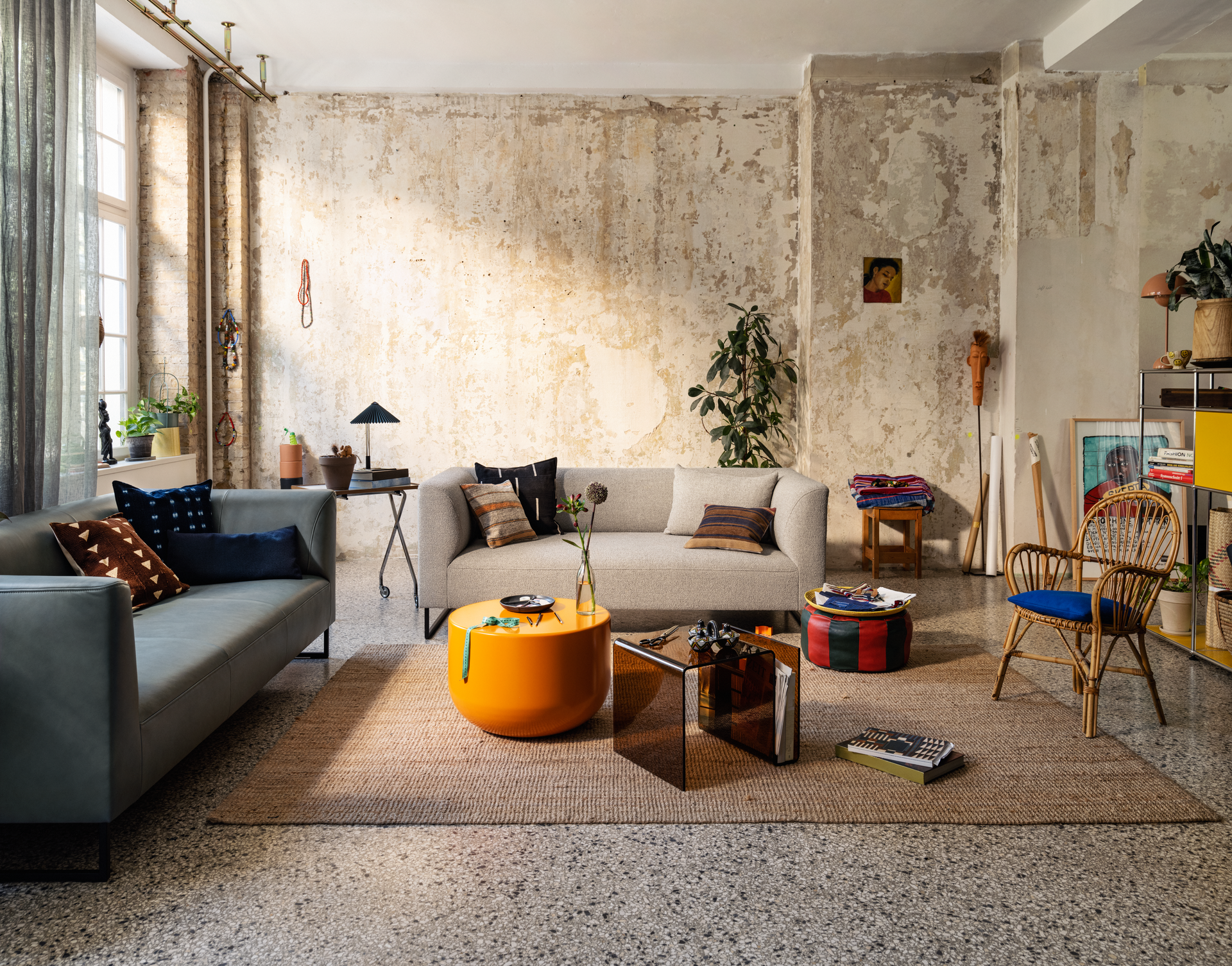 Wohnbereich mit grauem Sofa, orangefarbenem Couchtisch und Korbstuhl vor einer Wand im Used-Look