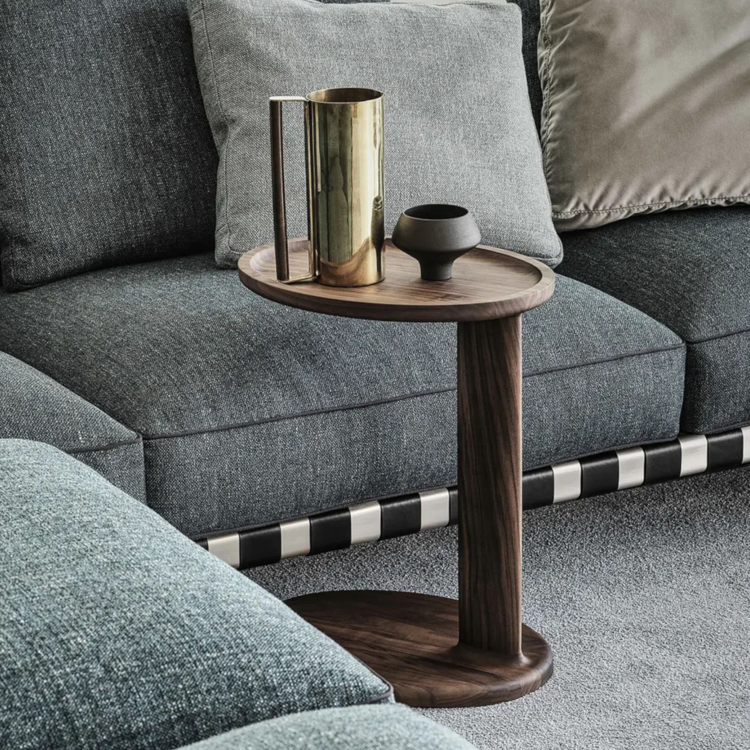 Zu sehen ist ein ovaler Beistelltisch in dunklem Holz neben einem grauen Stoffsofa. Auf dem Tisch steht ein glänzender Krug und eine kleine Vase.