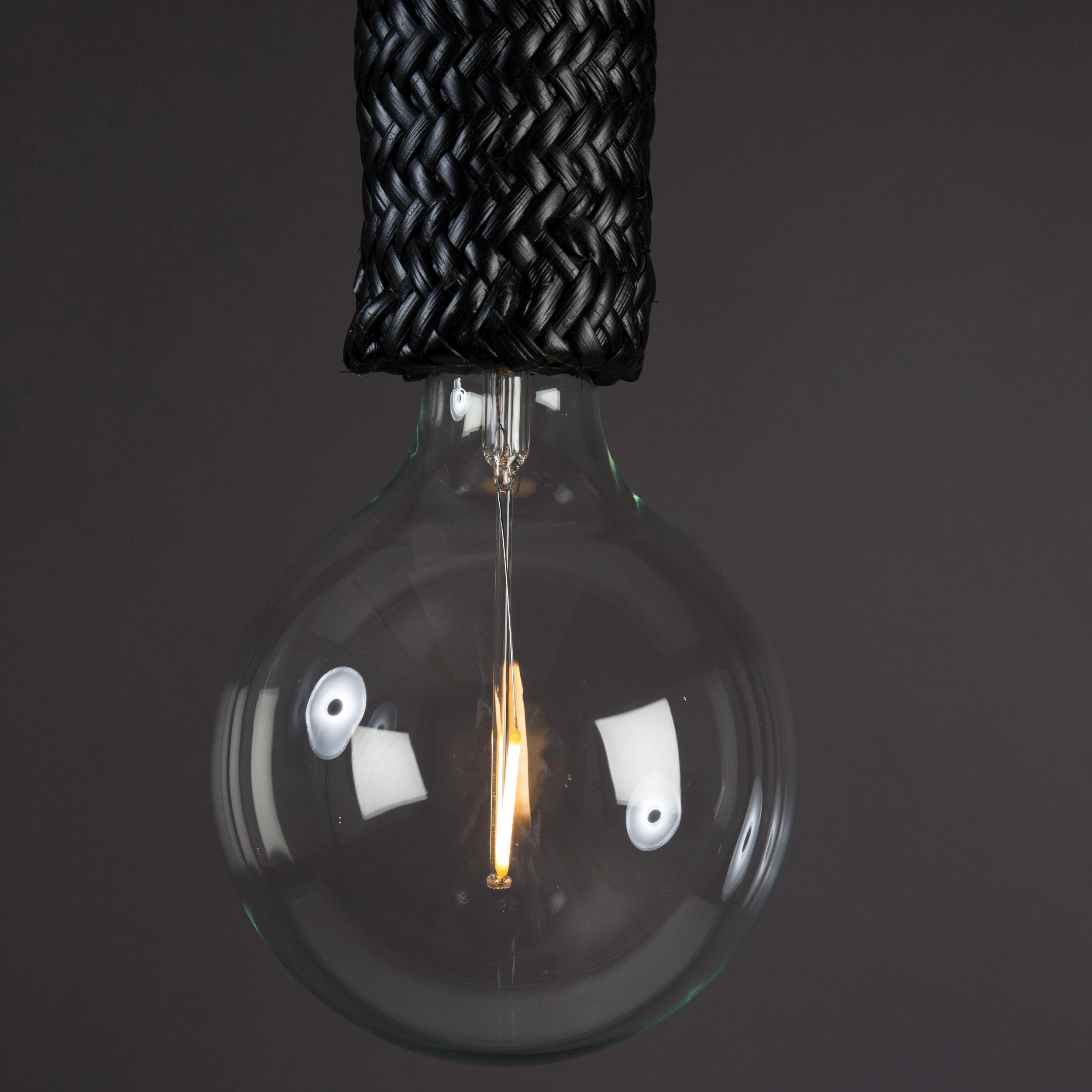 Nahaufnahme einer großen runden LED-Filament-Glühbirne an einer Hängelampe mit schwarz geflochtenem Rattan vor dunkelgrauem Hintergrund.
