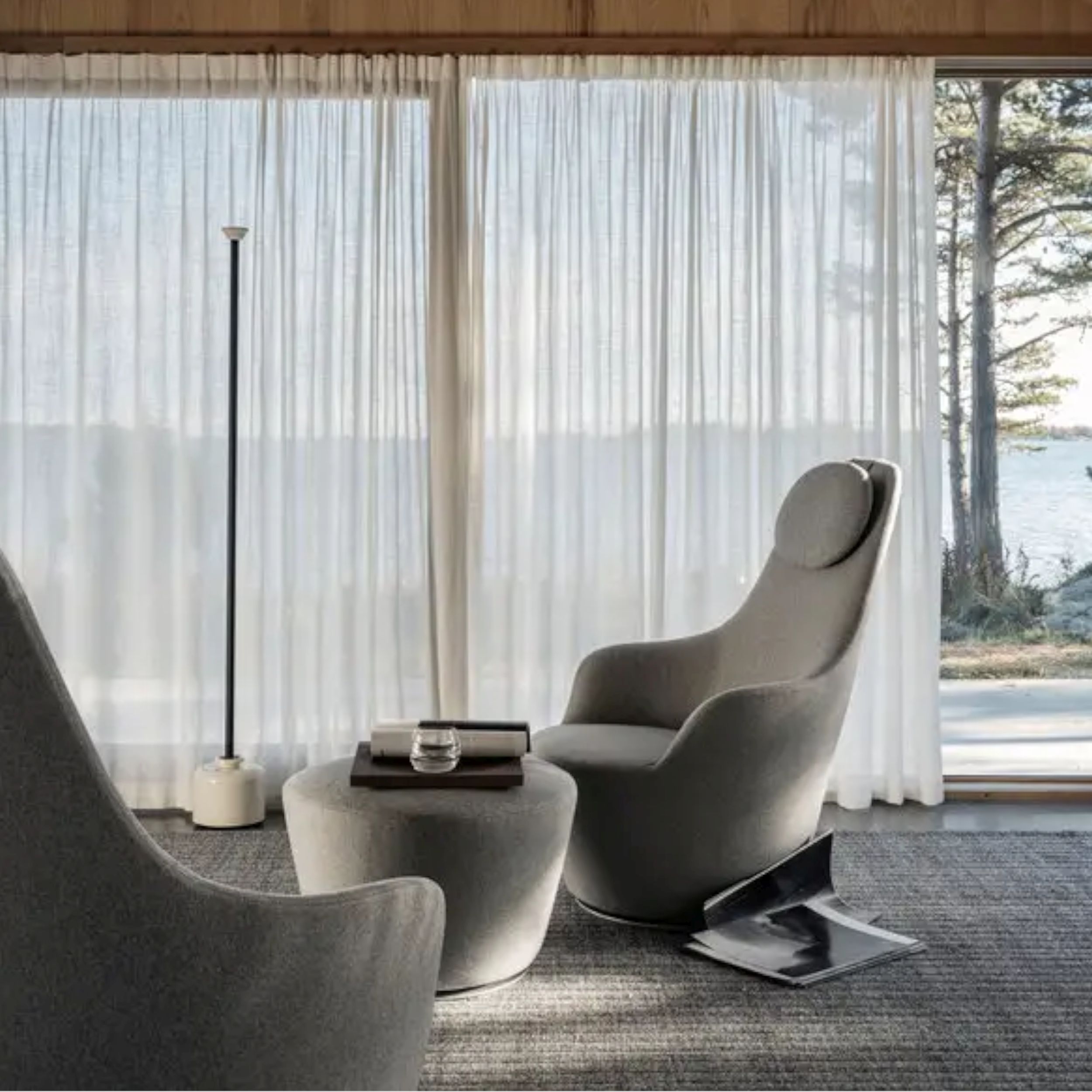 Elegantes Wohnzimmer mit großen Panoramafenstern; zentral im Bild steht der mit grauem Stoff bezogene Sessel Harbor von B&B Italia mit passendem Fußhocker auf einem grauen Teppich. 