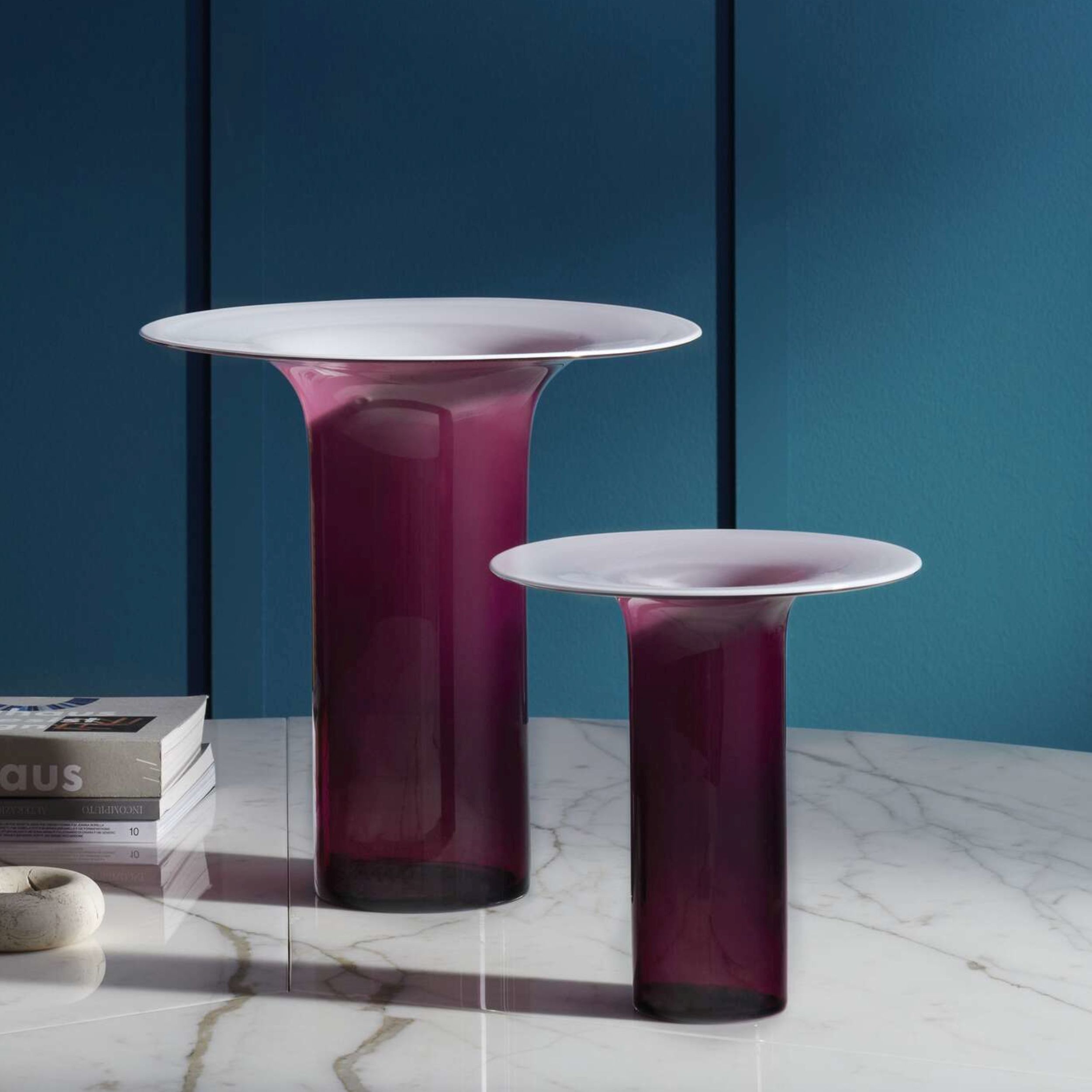 Zwei amethystfarbene Glasvasen, Modell Silvan von Cassina, stehen auf einem weißem Marmortisch vor einer blaugrünen Wand.