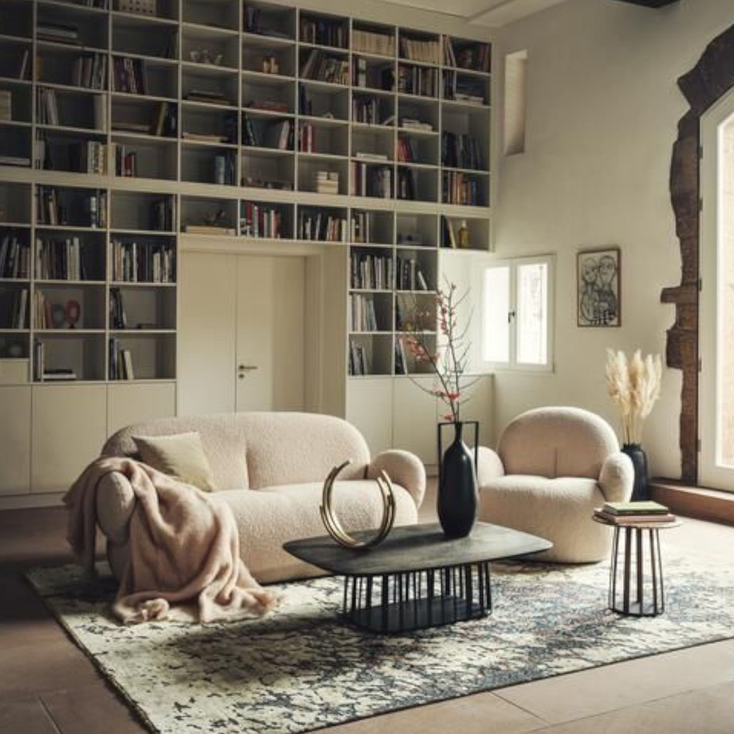 Vor einem großen, wandfüllenden Bücherregal steht eine weiße Polstergruppe bestehend aus Sofa und Sessel; ergänzt wird das Ensemble durch einen Couchtisch und einen Beistelltisch in dunklem Holz sowie einen gemusterten Teppich.