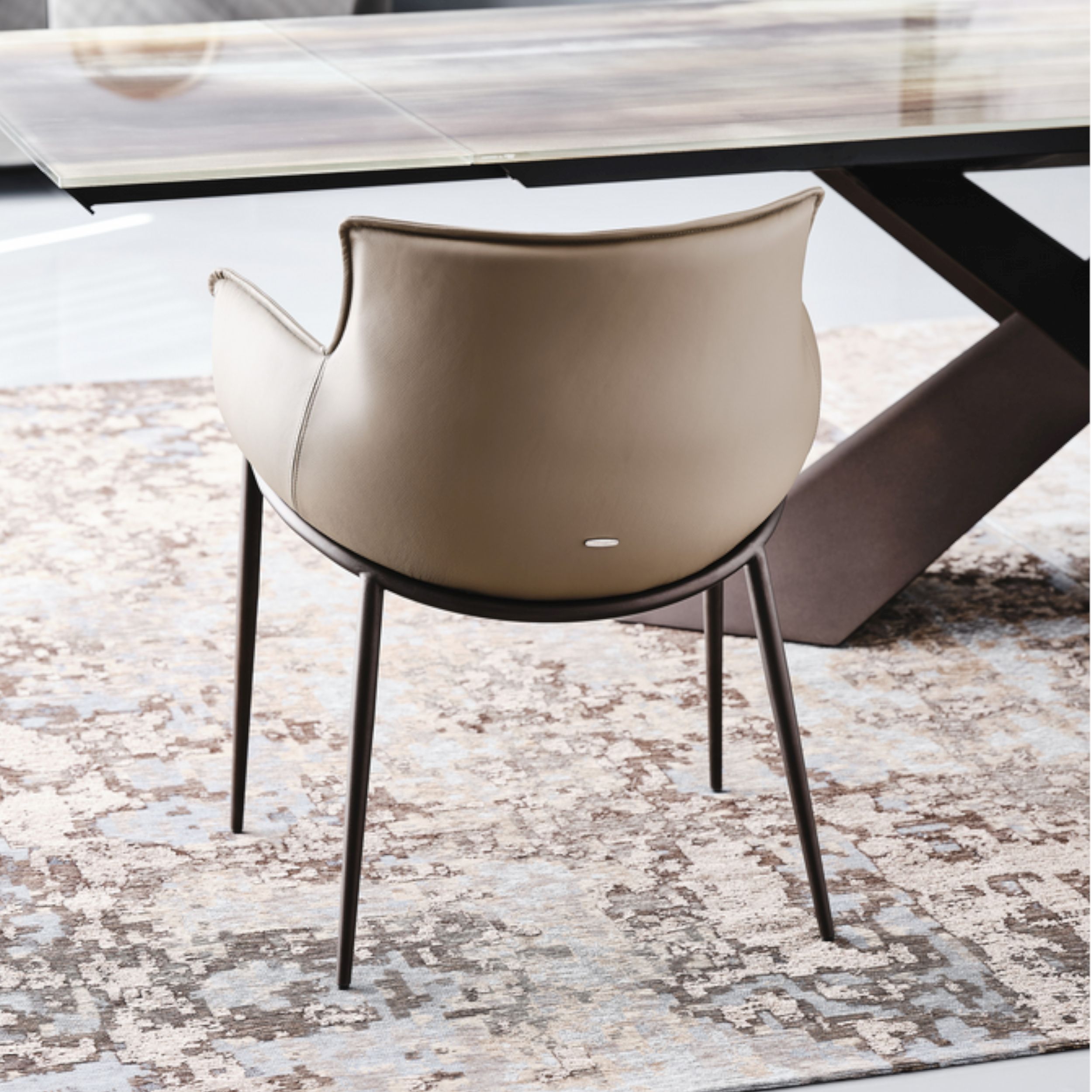 Ein eleganter beigefarbener Lederstuhl, Modell Rhonda von Cattelan Italia, in rückseitiger Ansicht vor einem Esstisch; Stuhl und Tisch stehen auf einem hellen, gemusterten Teppich.