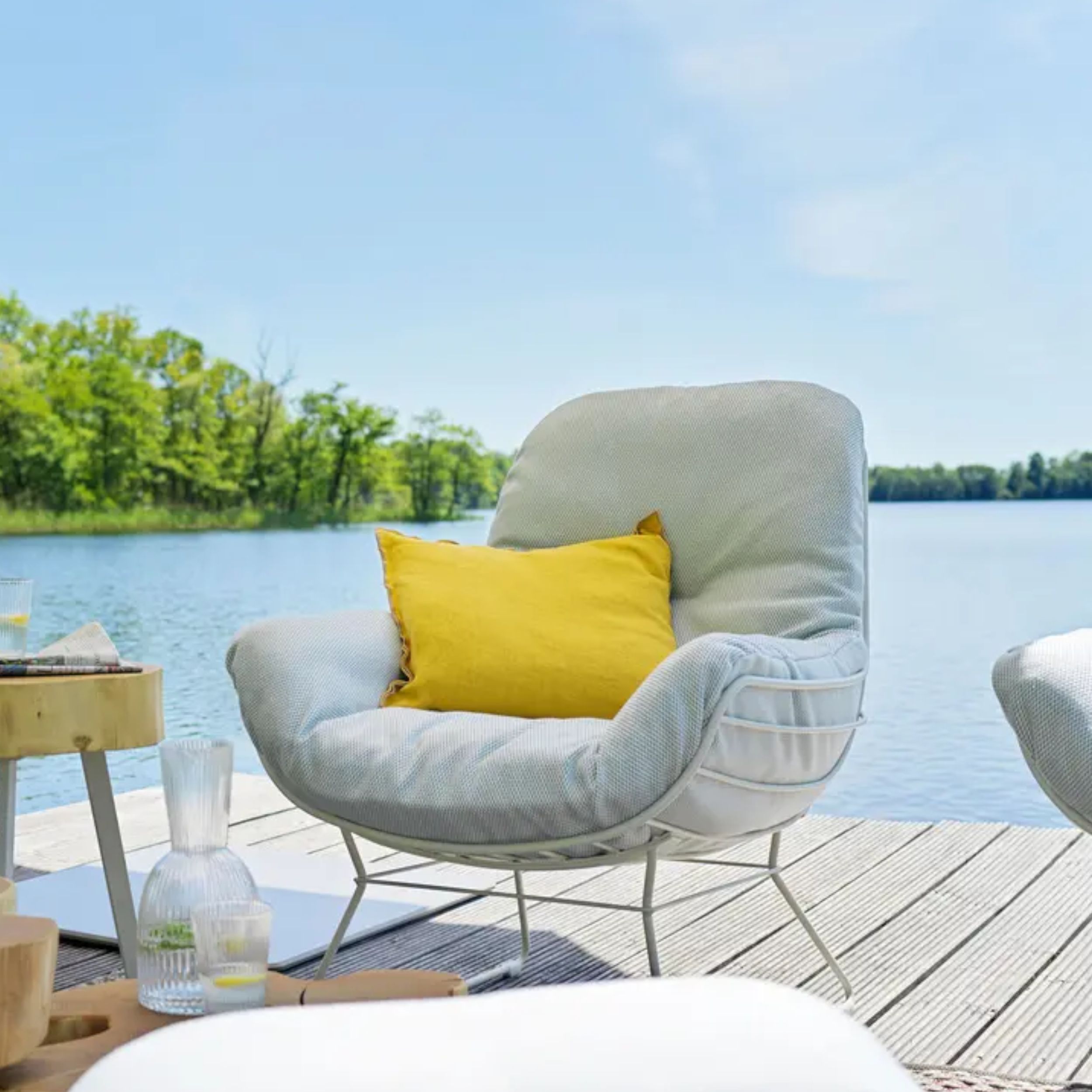 Zu sehen ist ein gemütlicher, heller Outdoorsessel auf einem Holzdeck vor einem See; im Sessel liegt ein gelbes Kissen.