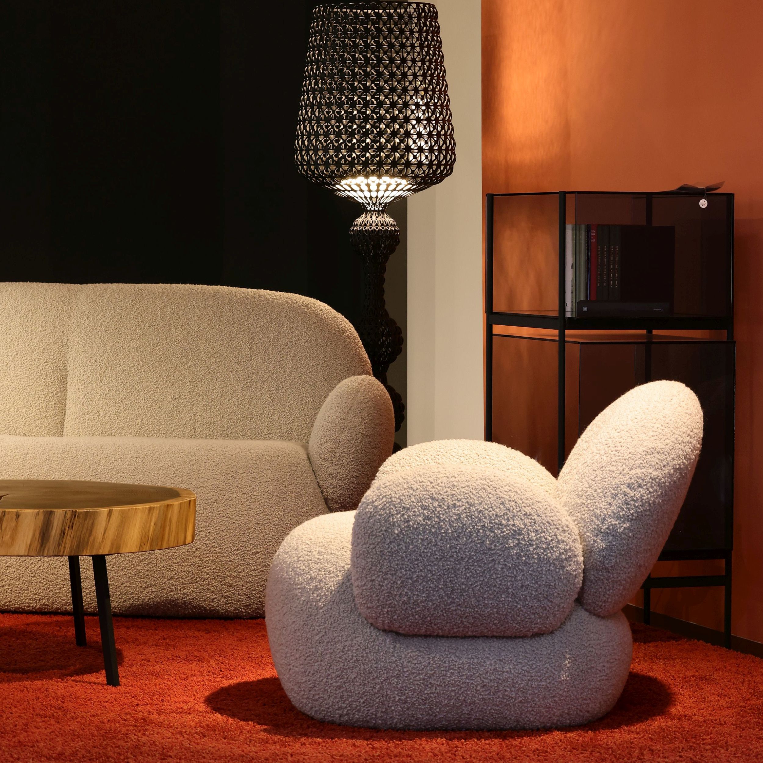 Zu sehen sind ein rundliches, weißes Sofa und ein passender Sessel vor einer Wand in rot und schwarz; die Möbel stehen auf einem roten Teppich und werden ergänzt durch einen Couchtisch, eine Stehleuchte und eine Vitrine.