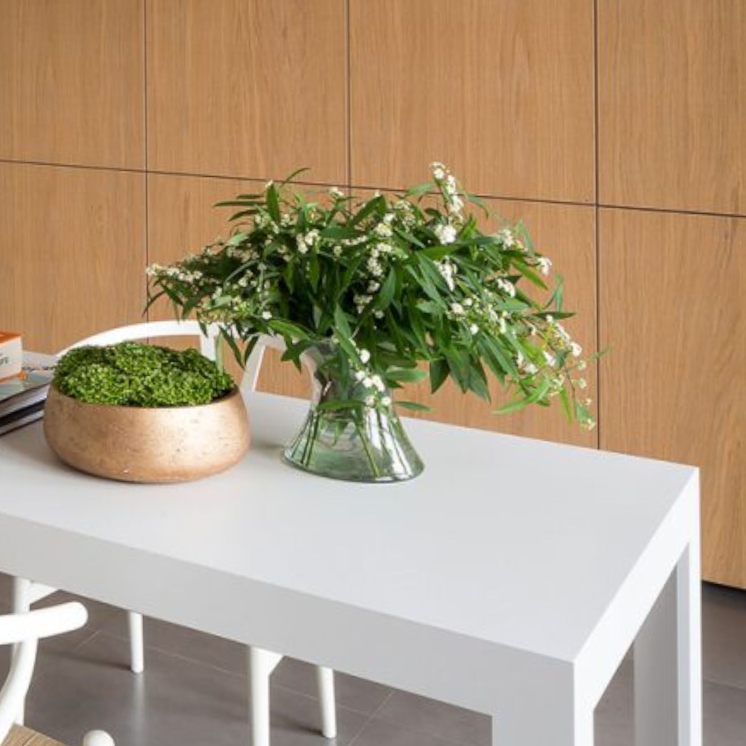 Zu sehen ist ein schlichter grauer Esstisch vor einer Wand mit Holzfronten, auf dem Tisch steht eine Vase mit einem grünen Strauß.