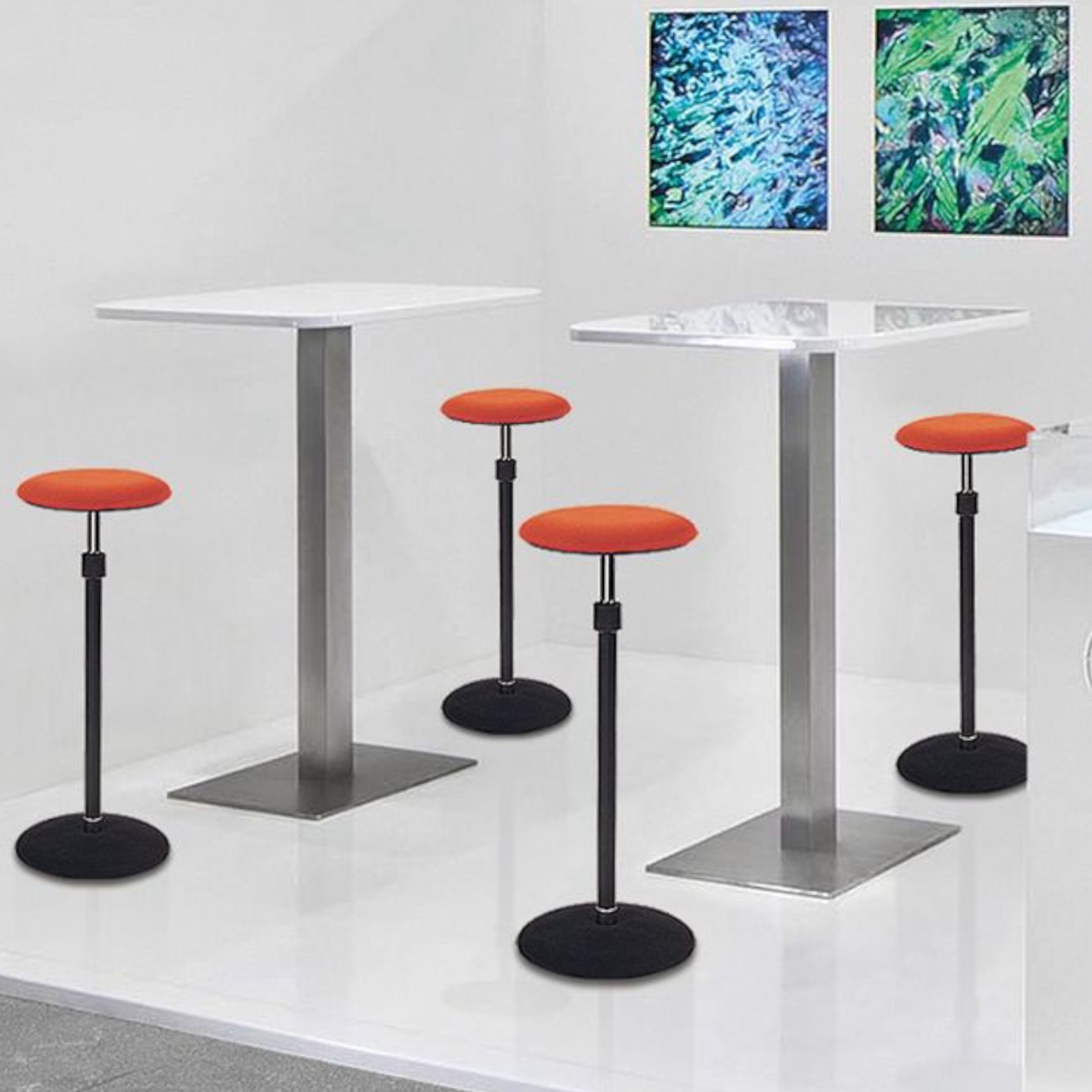 In einem in  weiß gehaltenen Raum stehen zwei eckige Stehtische und vier Steh-Sitz-Hocker in orange und schwarz; rechts im Hintergrund hängen zwei grüne Bilder an der Wand.