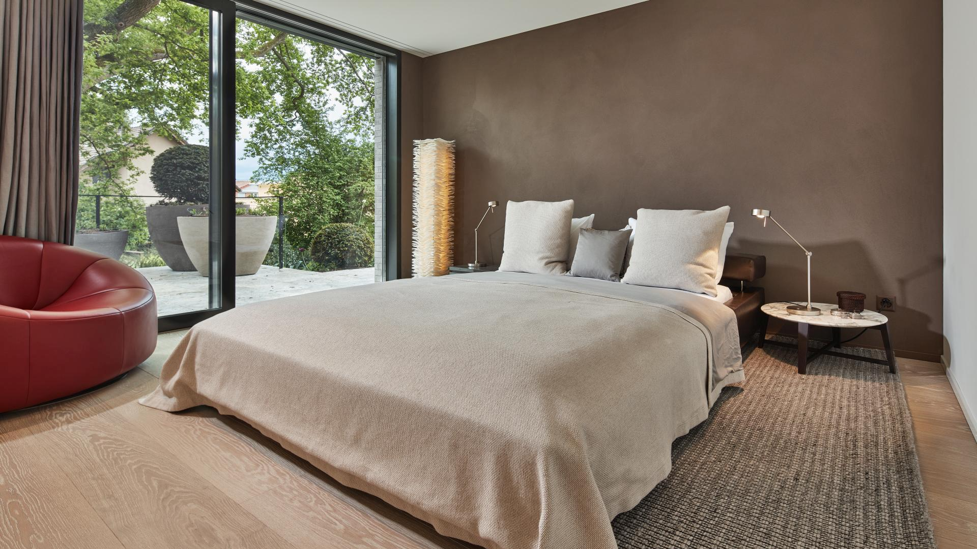 Schlafzimmer mit beigem Bett, großem Fenster und rotem Sessel