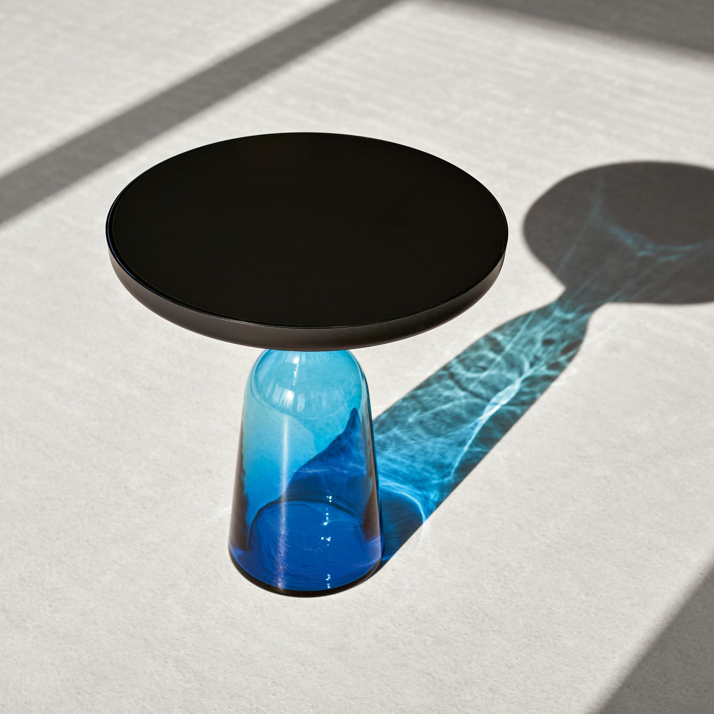 Ein runder Beistelltisch mit schwarzer Platte und blauem Glasfuß steht im Schlaglicht auf einem hellen Teppich; der Tischfuß wirft blaue Schatten auf den Boden.