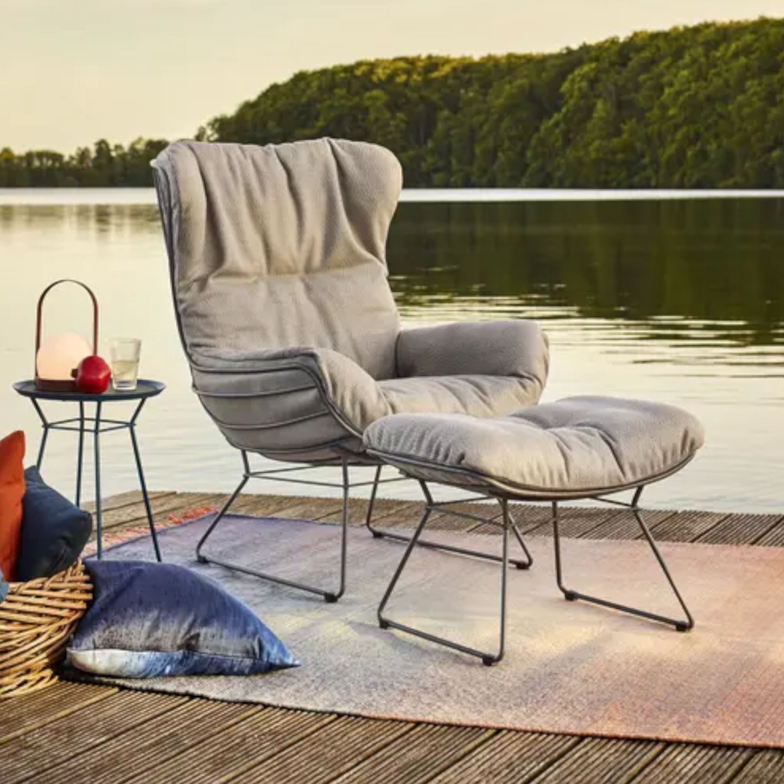 Zu sehen ist ein Loungesessel mit passendem Hocker und Beistelltisch auf einem Holzeck vor einem See, ergänzt durch einen roséfarbenen Teppich und ein paar Kissen.