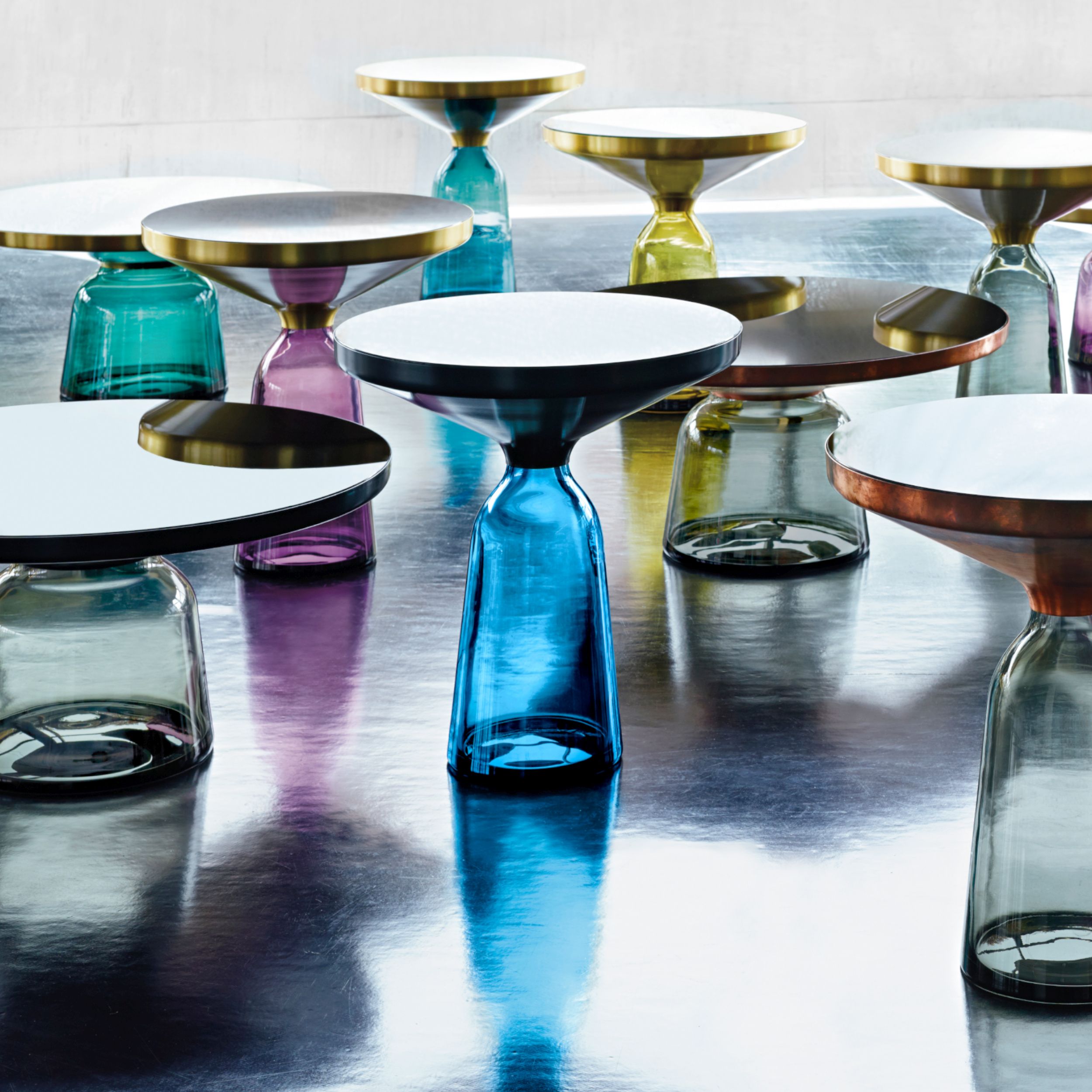 Eine Gruppe bunter Glastische in verschiedenen Größen und Farben steht auf einem glänzenden dunklen Boden.