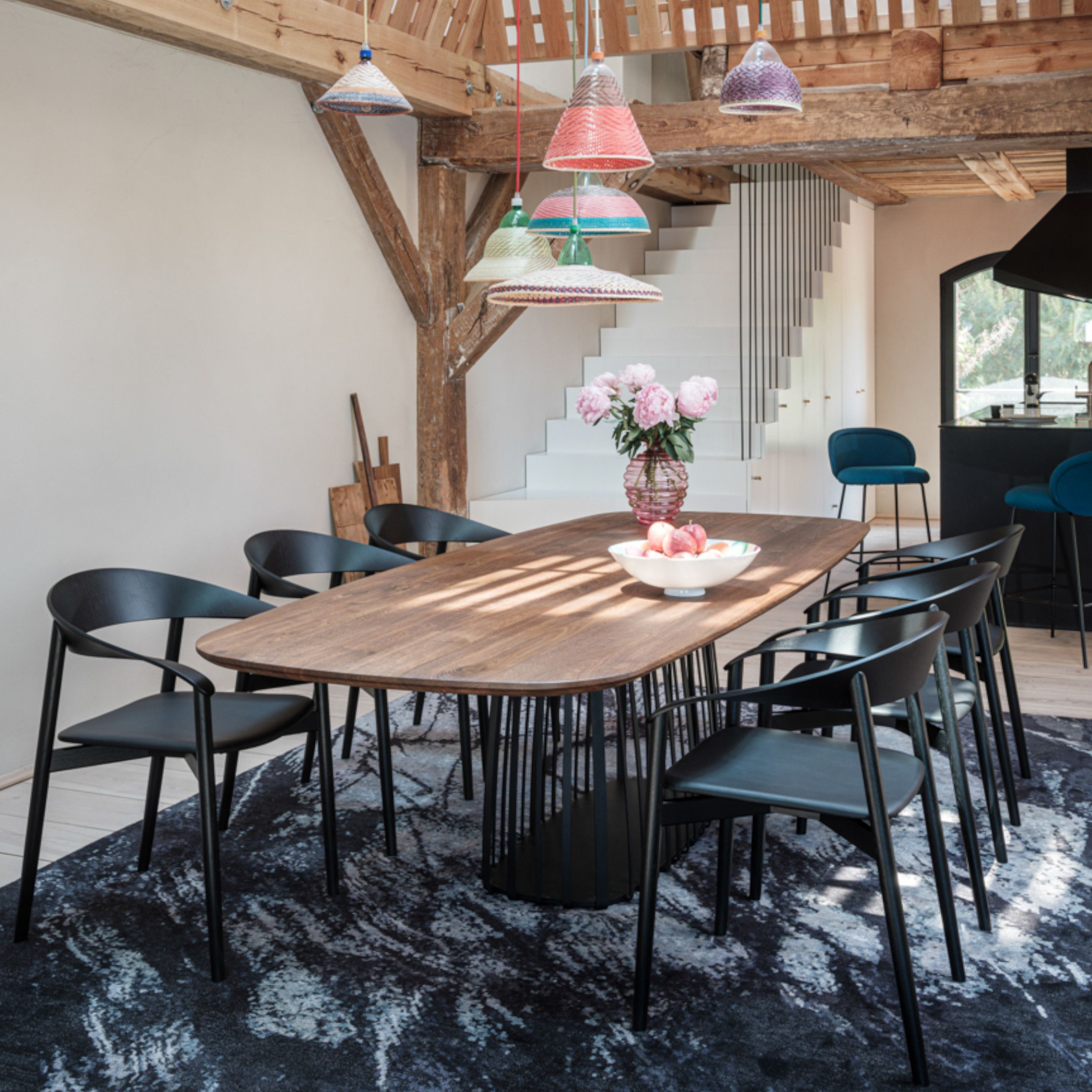 Zu sehen ist eine moderne Tischgruppe bestehend aus einem Holztisch und sechs schwarzen Stühlen auf einem blauen Teppich. Über dem Esstisch hängen mehrere bunte Leuchten, auf dem Tisch stehen Blumen; im Hintergrund sieht man eine weiße Treppe und Holzbalk
