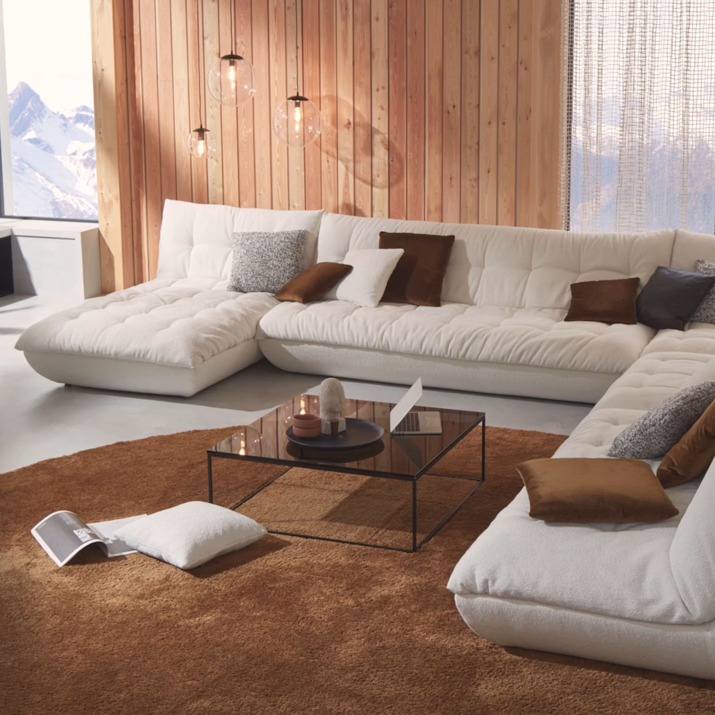 Das Bild zeigt einen gemütlichen Wohnraum mit einer großen Couchlandschaft in weißem Stoff. Das Sofa steht auf einem runden braunen Teppich, in der Mitte steht eine quadratischer Couchtisch mit Glasplatte.
