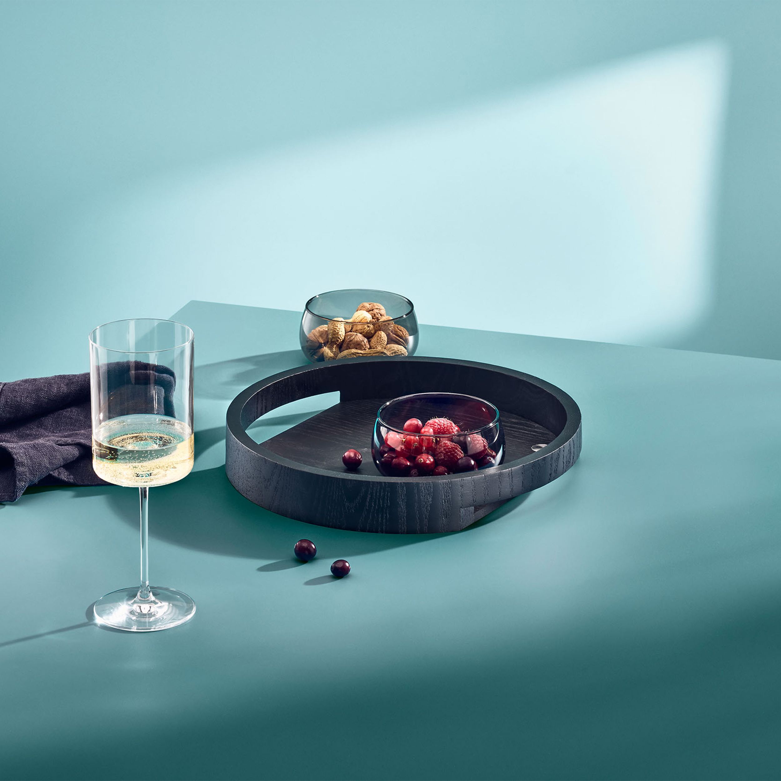 Rundes Servierbrett Style aus schwarzem Eschenholz mit sichtbarer Holzmaserung und integrierten Tragegriffen dekoriert auf einem
Tisch mit grüner Tischdecke und ein Glas Wein
sowie 2 kleine Schalen mit Snaks.