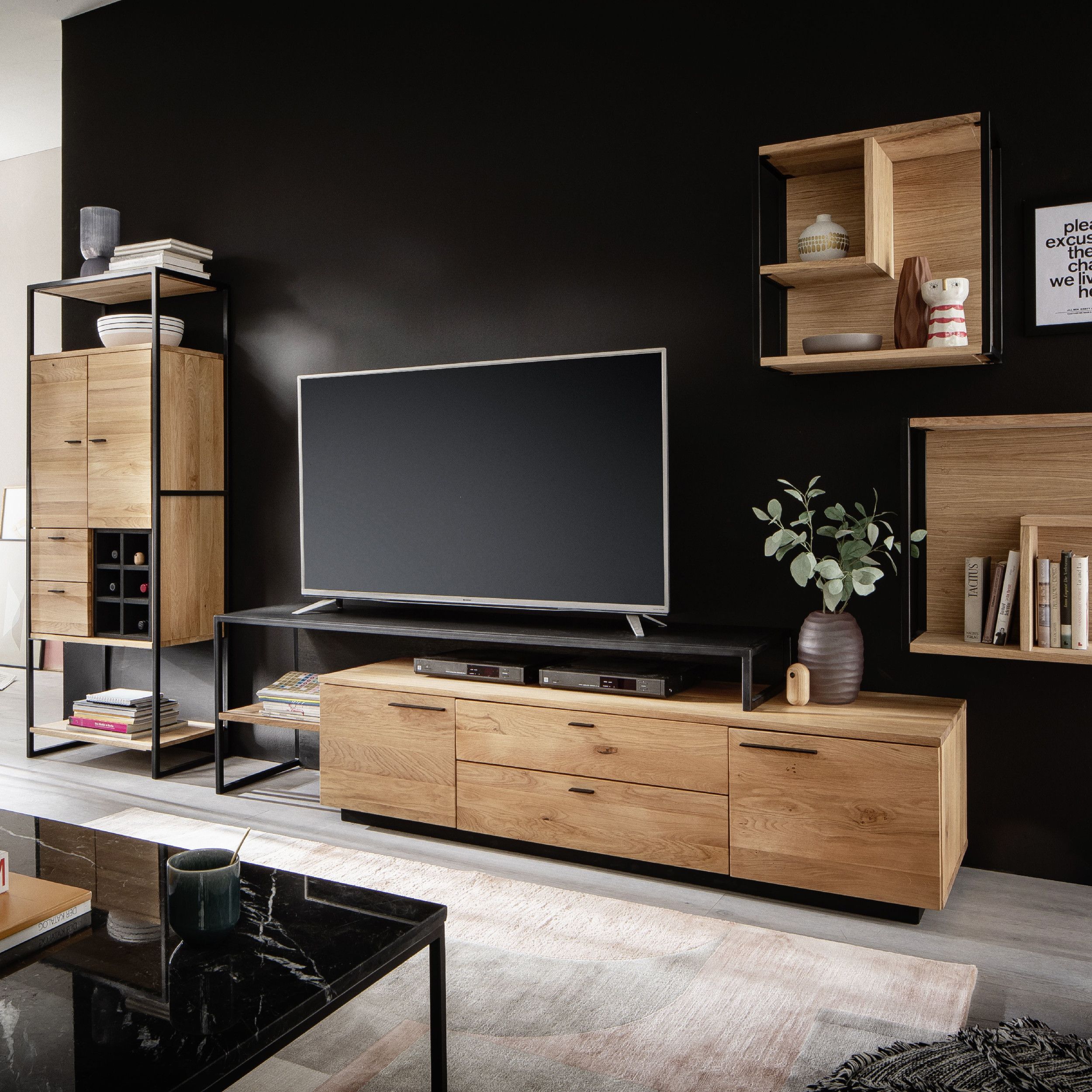 Einrichtungsbeispiel einer modernen Wohnwand aus hellem Eichenholz und schwarzem Metall vor einer schwarzen Wand, bestehend aus TV-Lowboard, hohem Regal, hängenden Wandregalen und passender Dekoration.