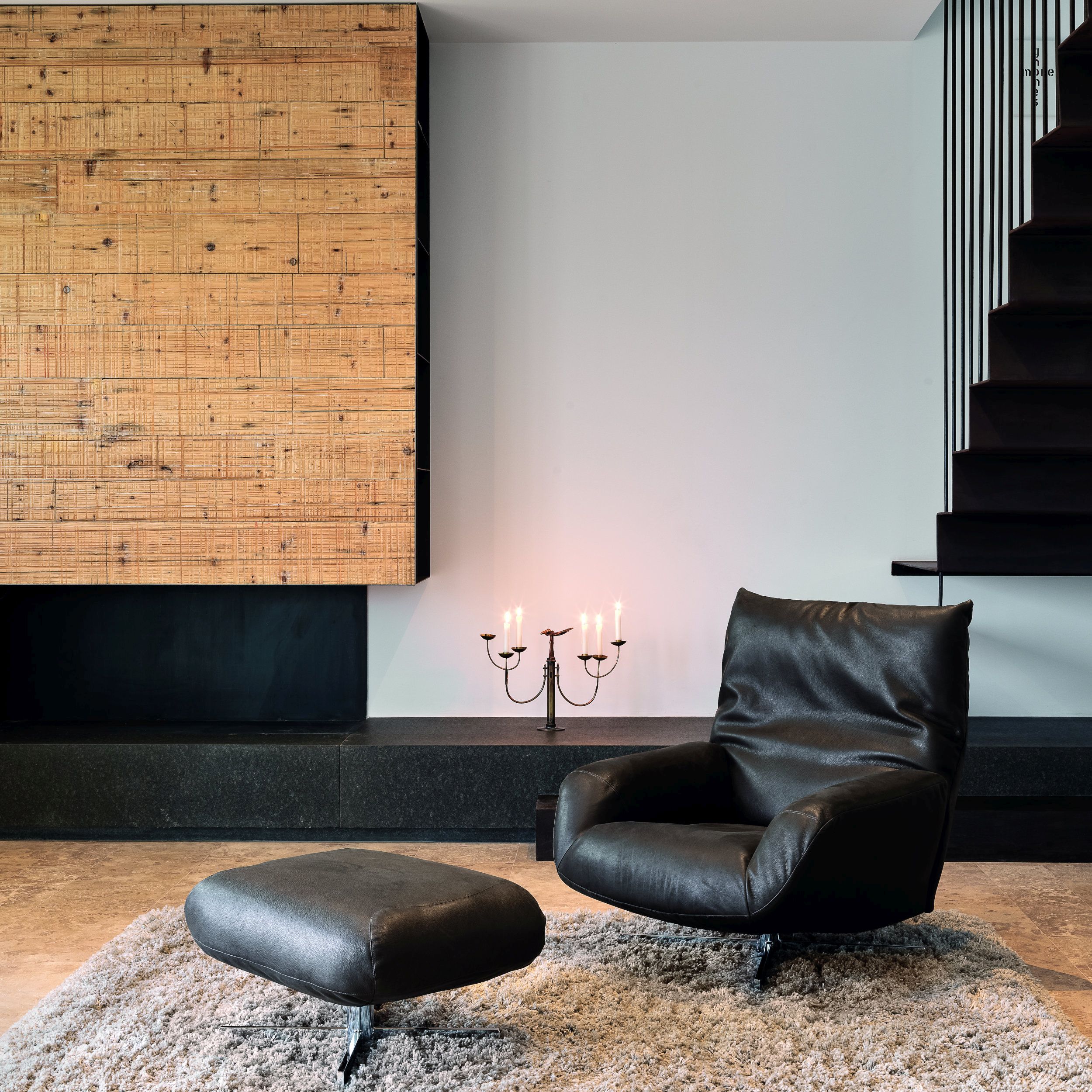 Das Bild zeigt ein minimalistisch-modernes Wohnzimmer mit einem schwarz gepolsterten Ledersessel samt Hocker auf einem grauen Teppich. Im Hintergrund sieht man einen schwarzen Kamin mit einer hellen Holzverkelidung, rechts erkennt man eine frei schwebende