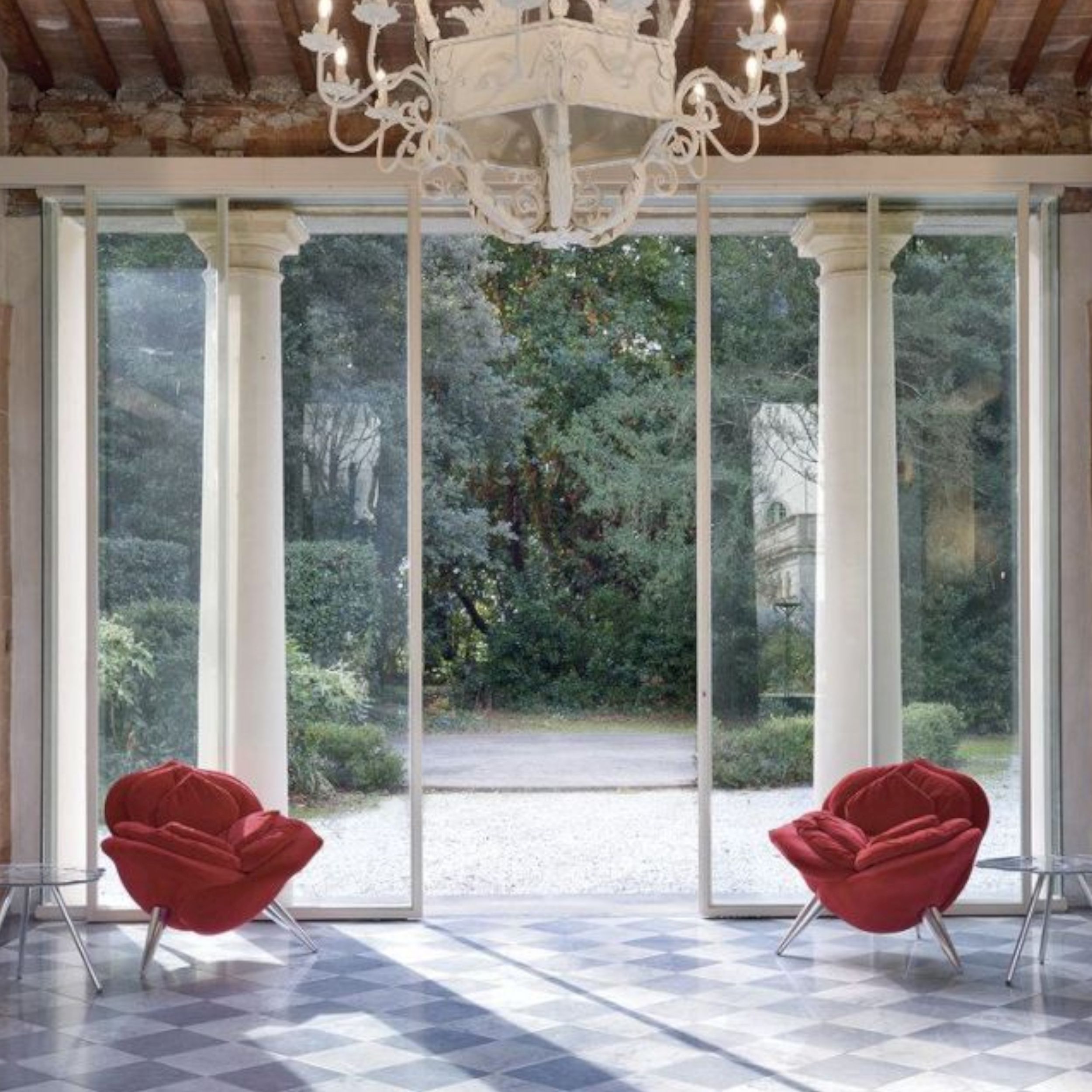 Das Bild zeigt den Blick aus einer klassischen Villa mit weißen Säulen hinaus in den Garten; der Ausgang wird flankiert von zwei roten Sesseln in Form von Rosenblüten.