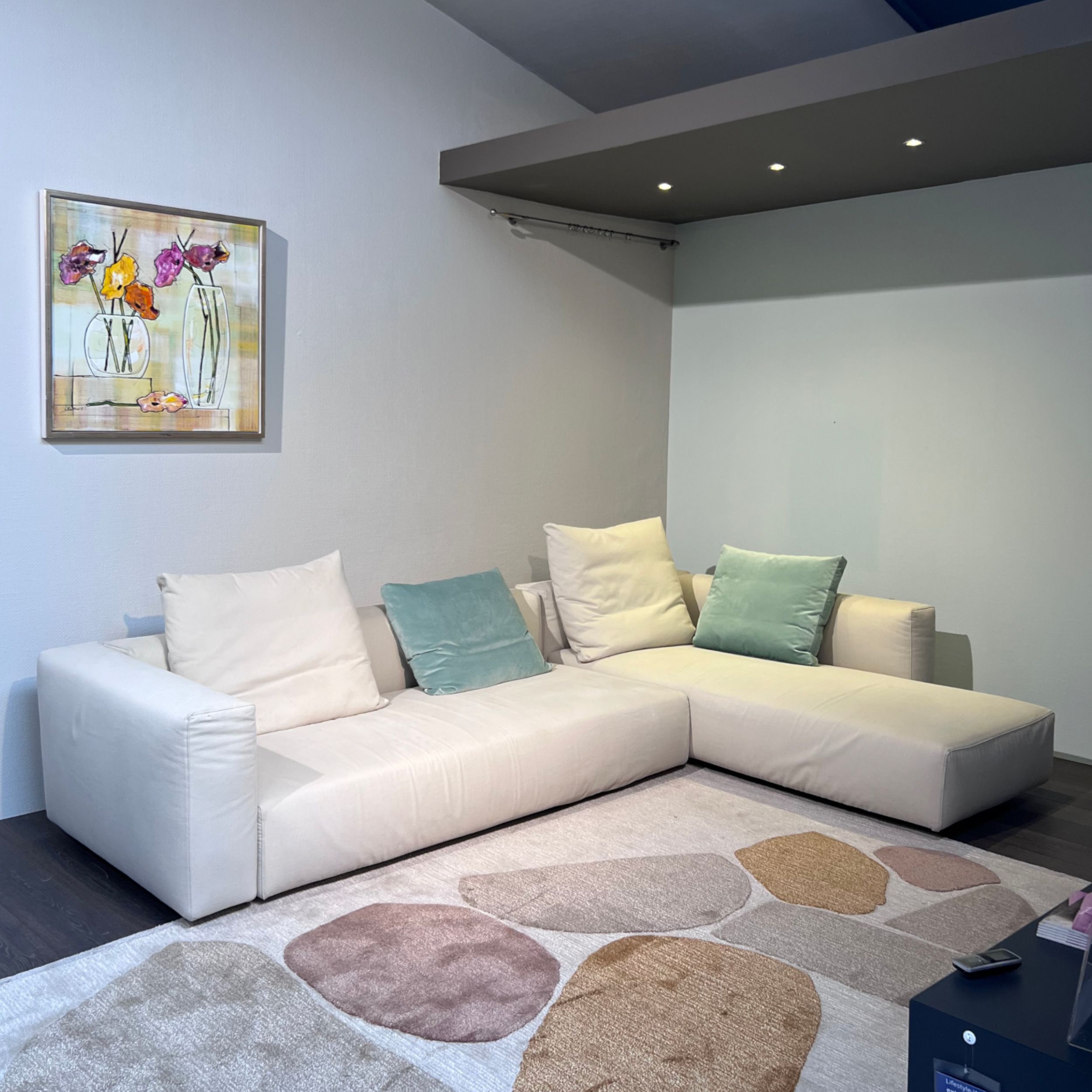 Zu sehen ist das Sofa Lario von Flexform in einer L-förmigen Kombination mit offen endenem Liegenelement rechts. Das Sofa ist mit naturfarbenem Stoff bezogen und steht auf einem dunklen Holzboden. Vor dem Sofa liegt ein heller Teppich mit Ornamenten in ro