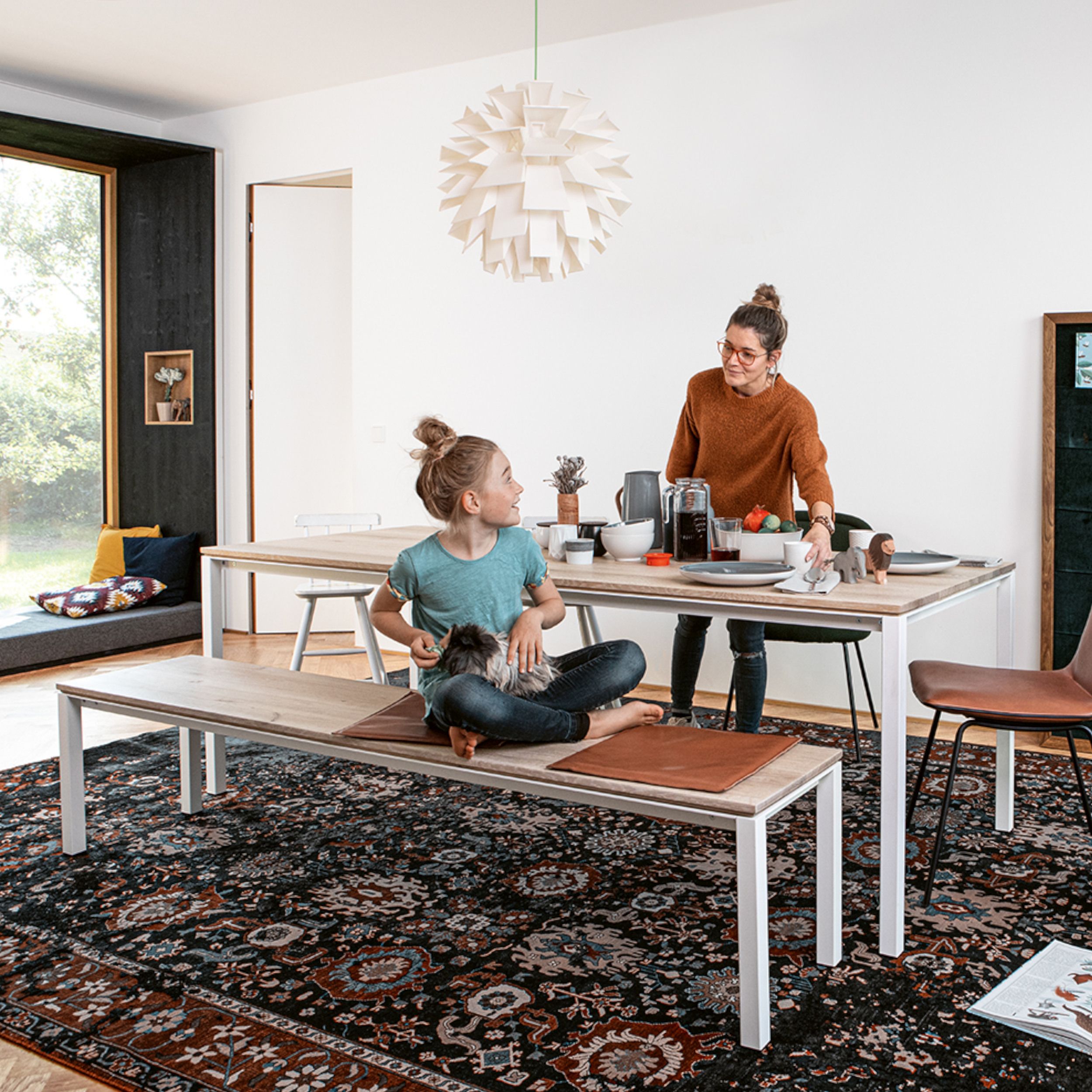 Das Bild zeigt ein  modernes Esszimmer mit zwei Personen, die sich am Tisch aufhalten. Auf dem Boden liegt eine großer Orientteppich, über dem Tisch hängt eine große runde Leuchte. Der Tisch ist gedeckt, auf der Bank daneben sitzt ein Kind.