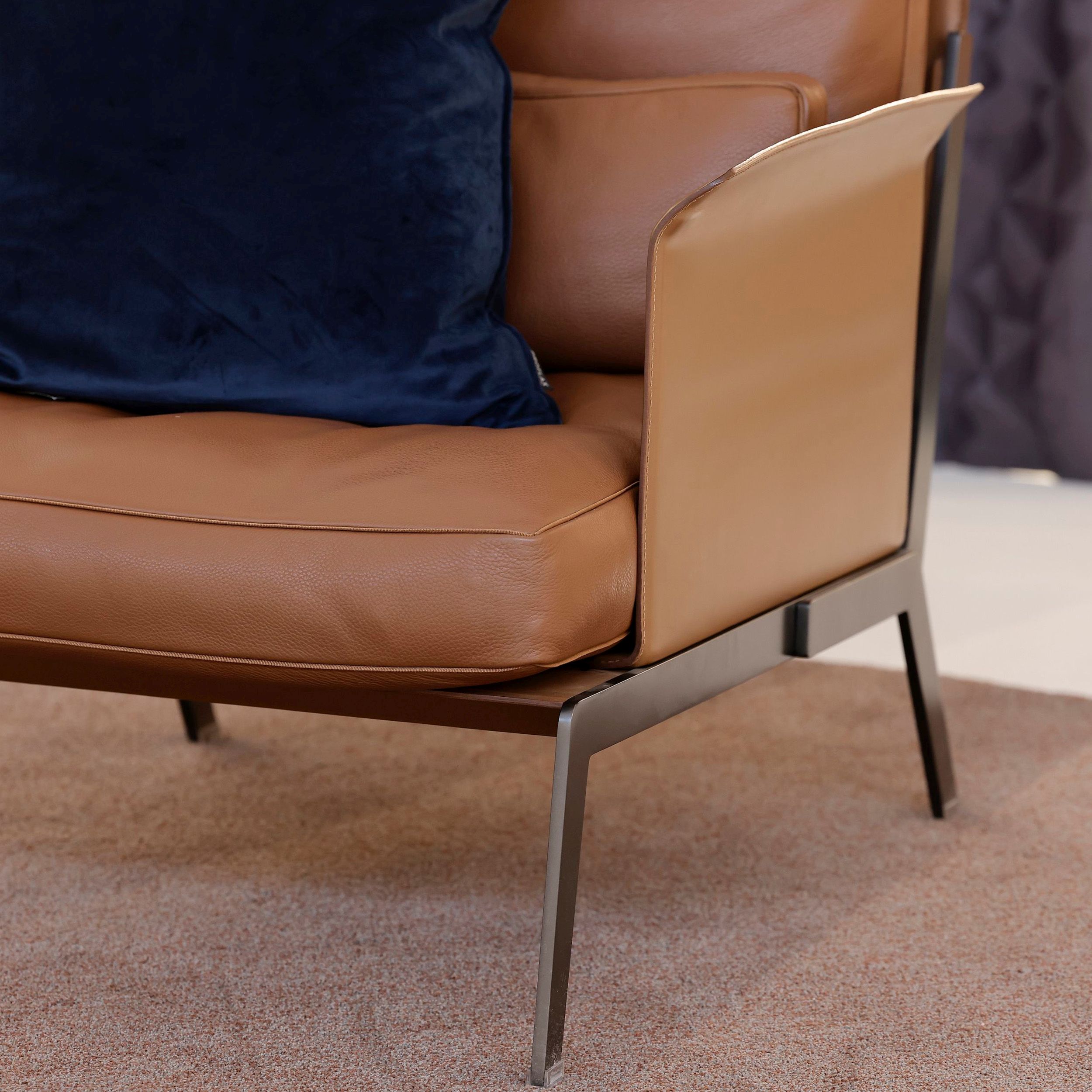 Das Bild zeigt eine Detailaufnahme des Sessel-Modells Happy Hour von Flexform: Man sieht die kernlederbezogene, gebogene Armlehne, das matte Flachstahlgestell und die aufliegenden Polsterkissen.