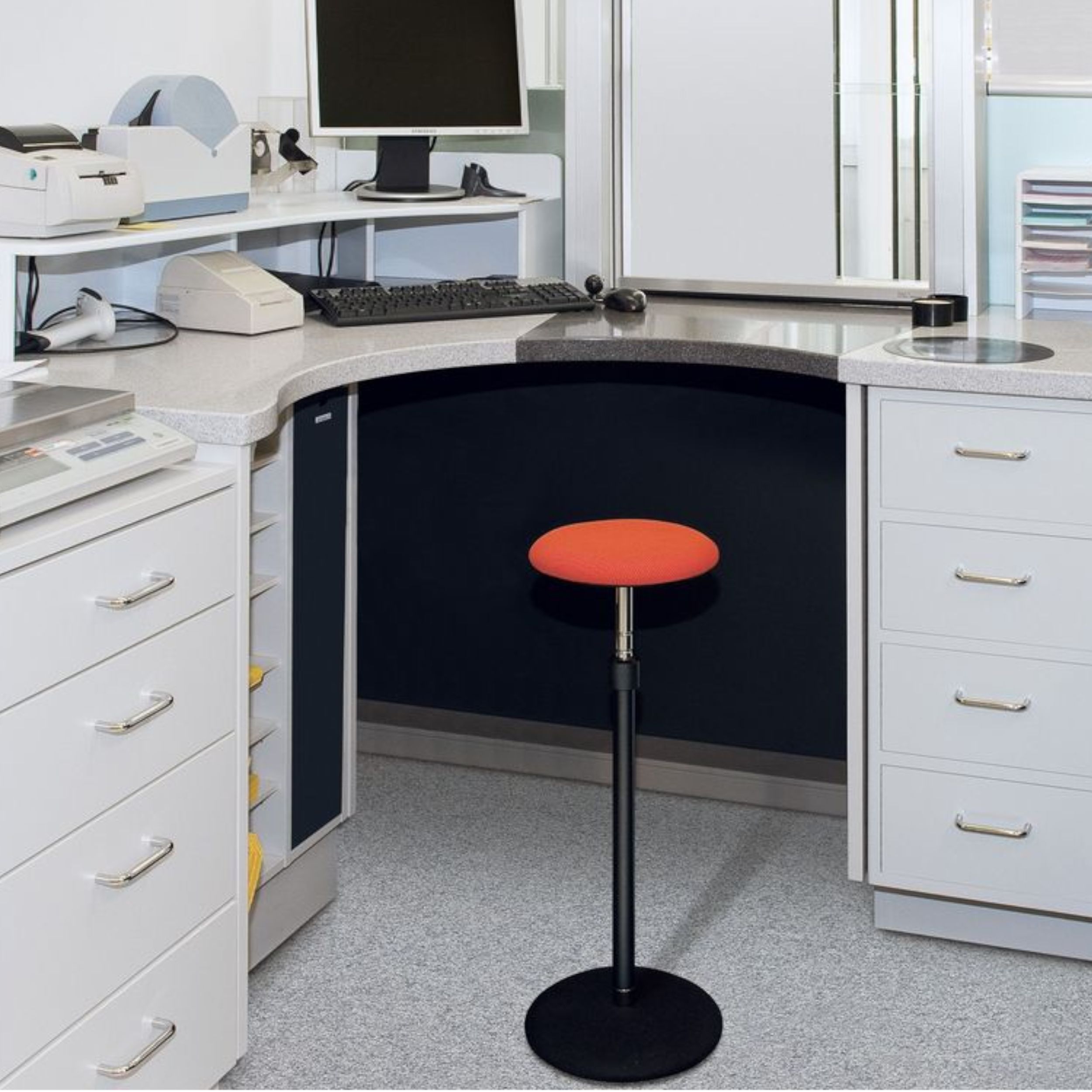 An einem Büro-Arbeitsplatz steht ein Steh-Sitz-Hocker mit orangefarbenem Sitzpolster und schwarzem Säulenfuß.