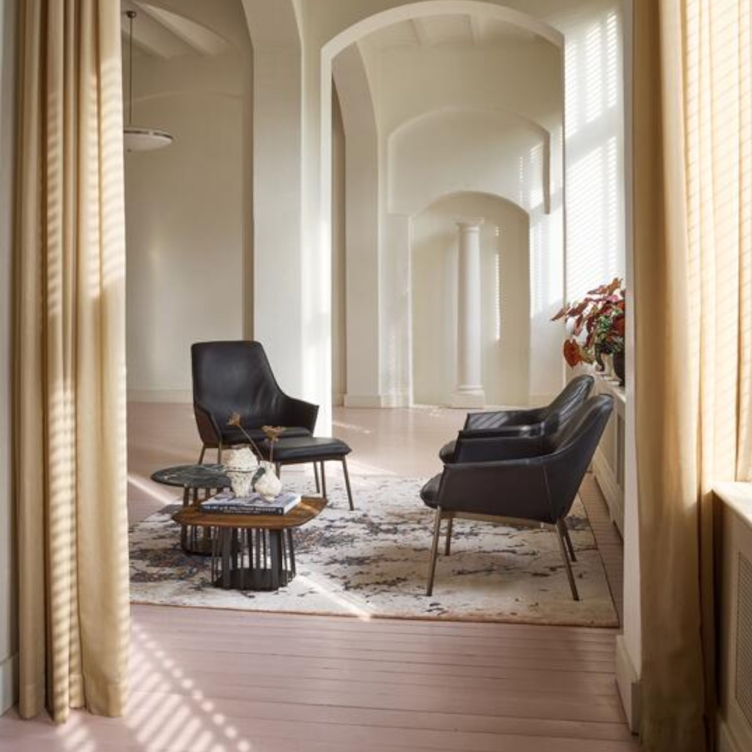 Zu sehen ist eine Sitzgruppe bestehen aus drei dunklen Ledersesseln und zwei Tischchen in einem eleganten hellen Raum mit hohen Decken. Die Sessel stehen auf einem gemusterten Teppich, im Vordergrund wird der Eingang in den Raum von hellbraunen Stoffvorhä