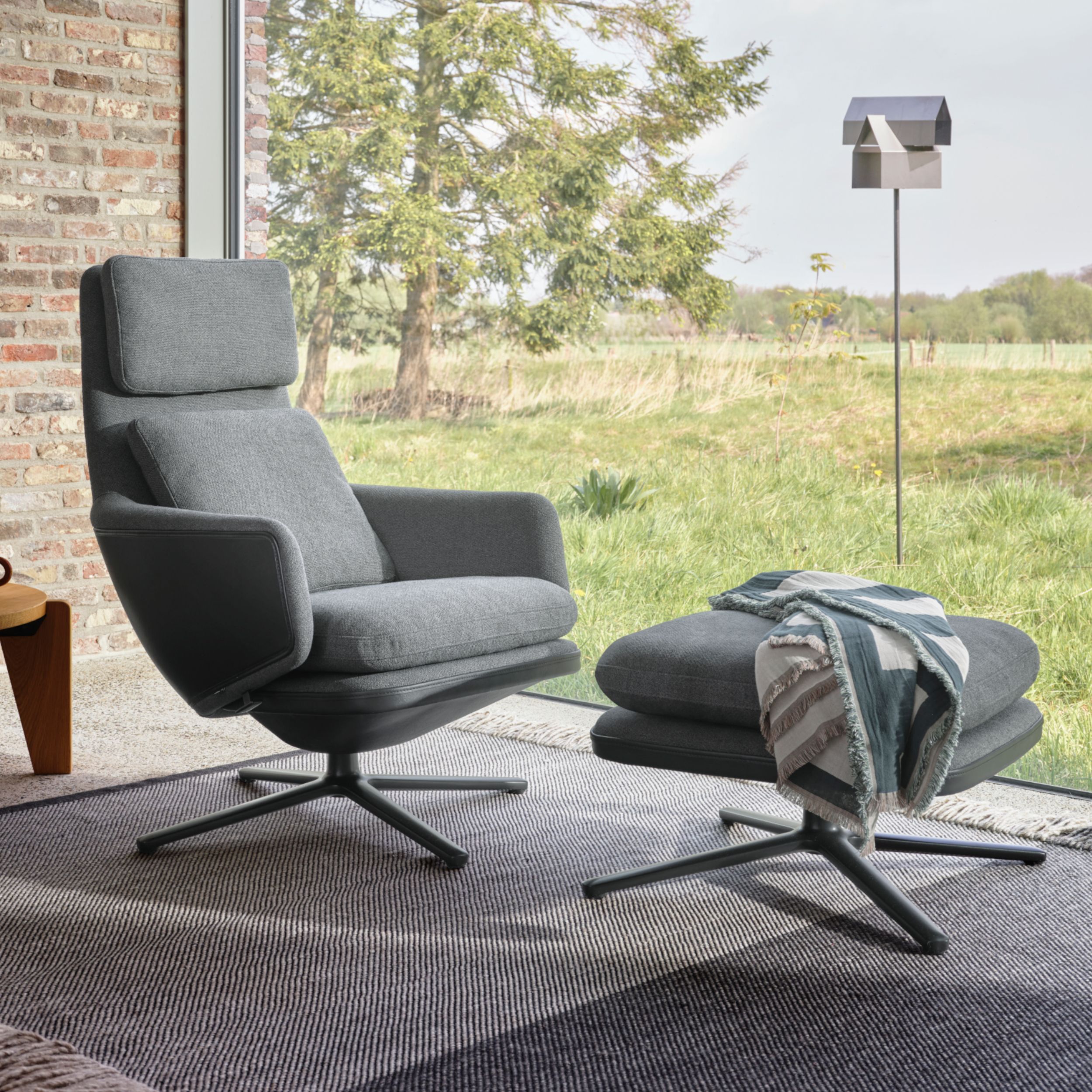 Auf dem Bild zu sehen ist der Relaxsesssel Grand Relax von Vitra mit dem dazugehörigem Hocker Ottoman in einer Kombination aus grauem Stoff und schwarzem Leder. Der Sessel steht in einem modernen Raum mit Backsteinwand vor einem Fenster mit Ausblick auf e