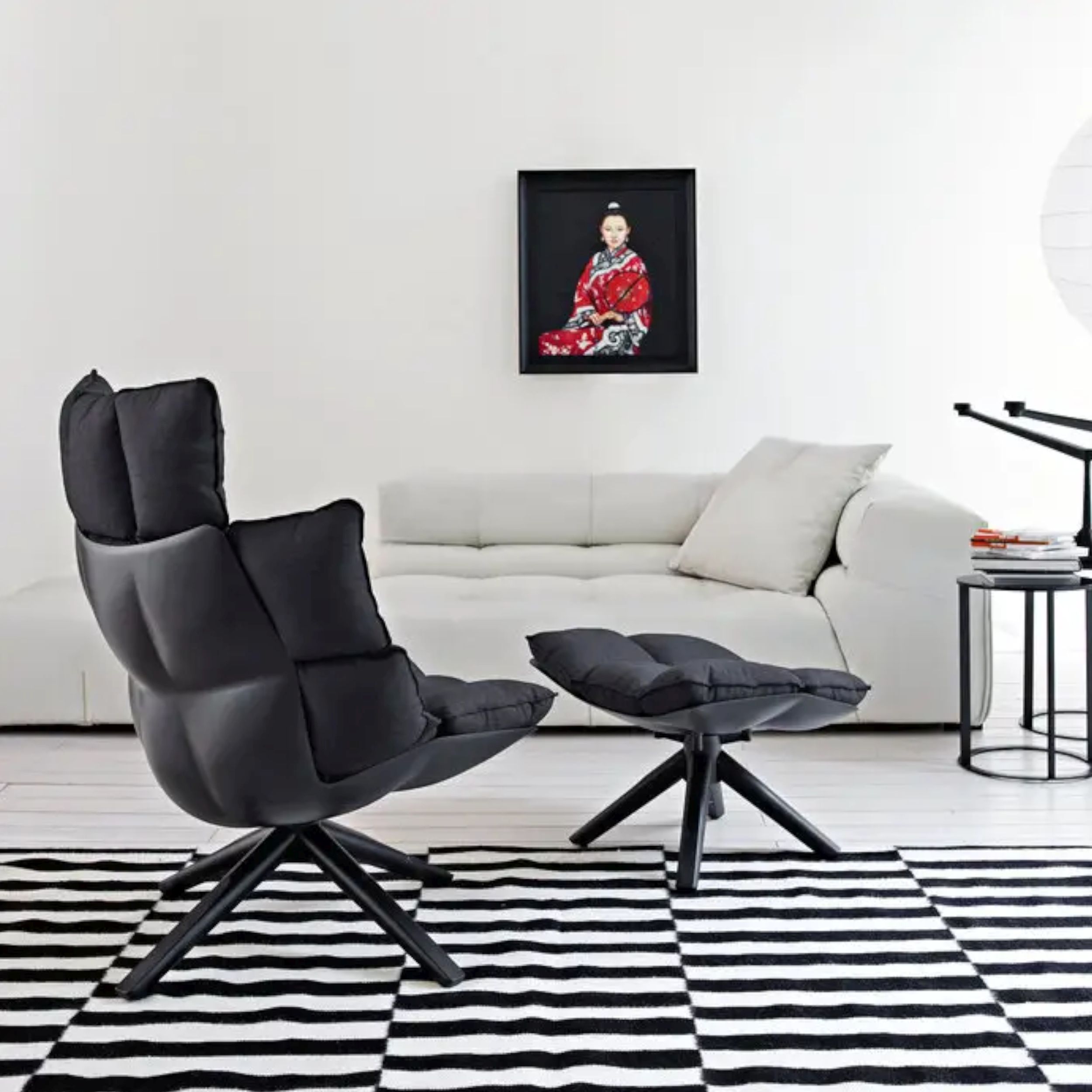 Sessel Husk von B&B Italia in schwarz mit Hocker in einem modernen Wohnraum auf einem schwarz-weiß- gemusterten Teppich; im Hintergund zu ein weißes Sofa unter einem schwarz-roten Bild.