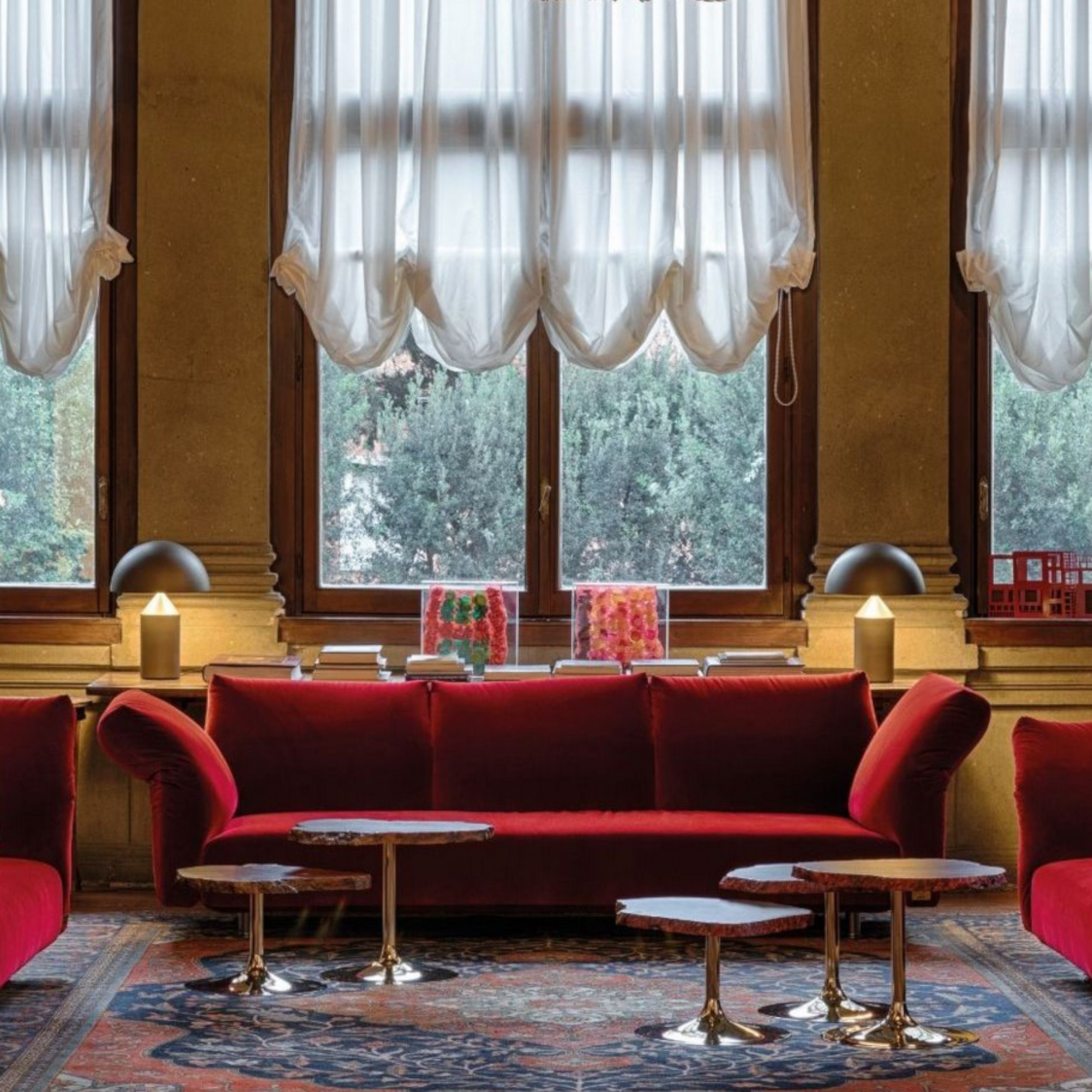 Ein rotes Stoffsofa steht in einem opulent gestalteten Raum, ergänzt durch mattgoldene Tischleuchten und eine Gruppe organischer Couchtische auf einem Orient-Teppich.