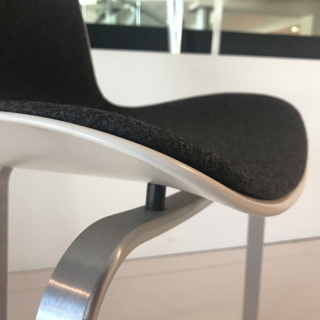 Ein dreibeiniger Stuhl, Modell PK8 von Fritz Hansen, in Detailaufnahme.
Zu sehen ist von der Seite die weiße Kunststoff-Sitzschale mit grauer Polsterung und darunter das dreibeinige Untergestell aus matt gebürstetem Aluminium.
