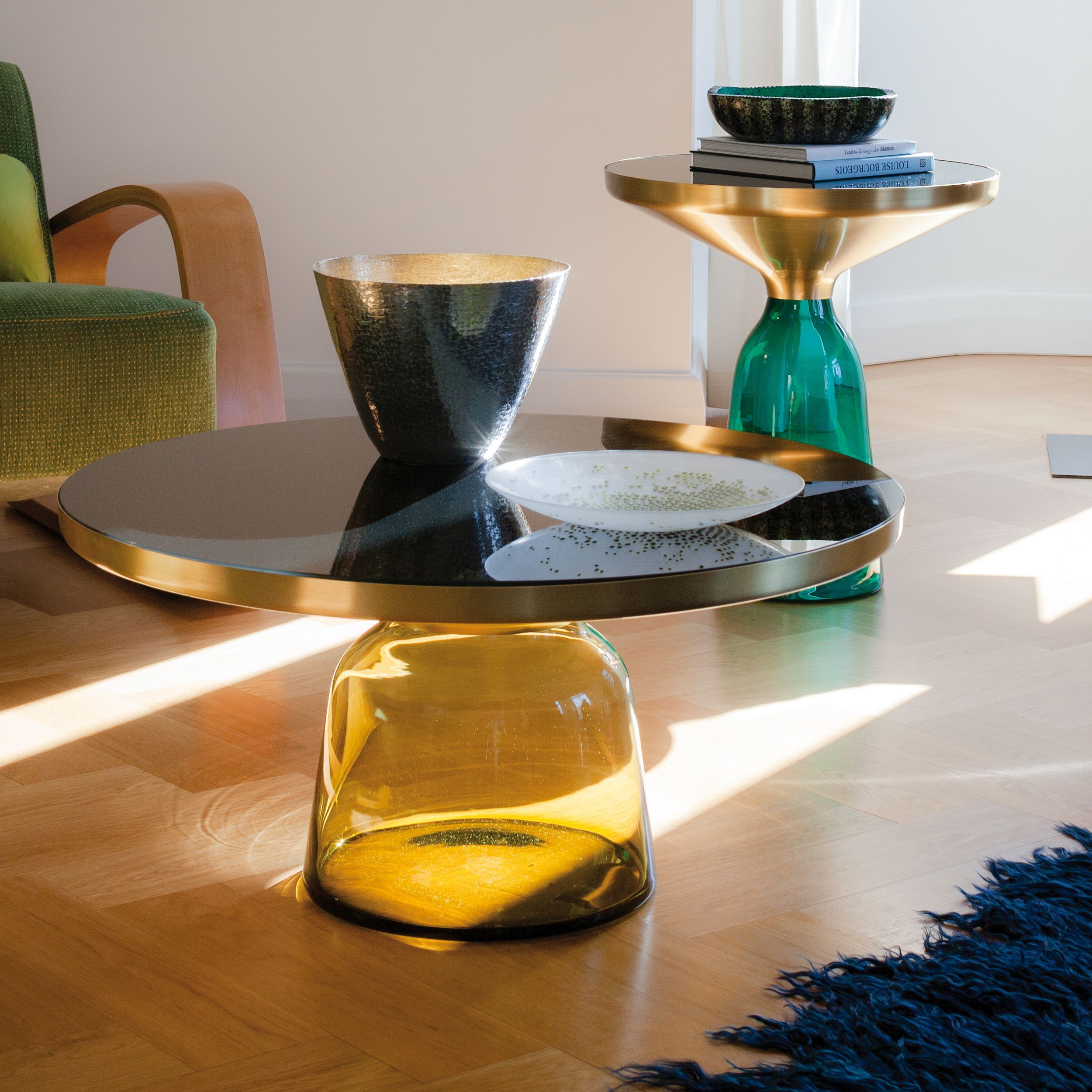 Zwei elegante Couchtische mit farbigem Glasfuß und Messingdetails stehen auf einem Parkettfußboden, im Hintergrund links ist ein grüner Sessel zu sehen.