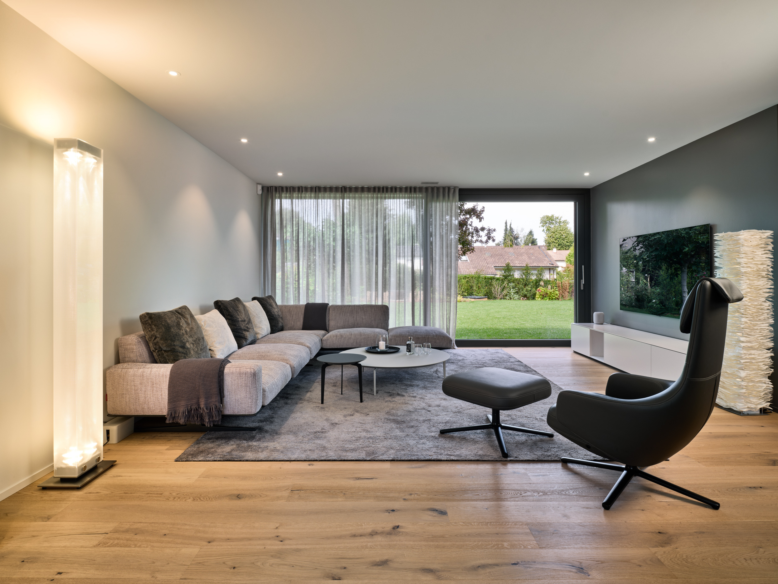 Wohnzimmer mit Ecksofa, Stehleuchte, schwarzem Sessel und Blick in den Garten