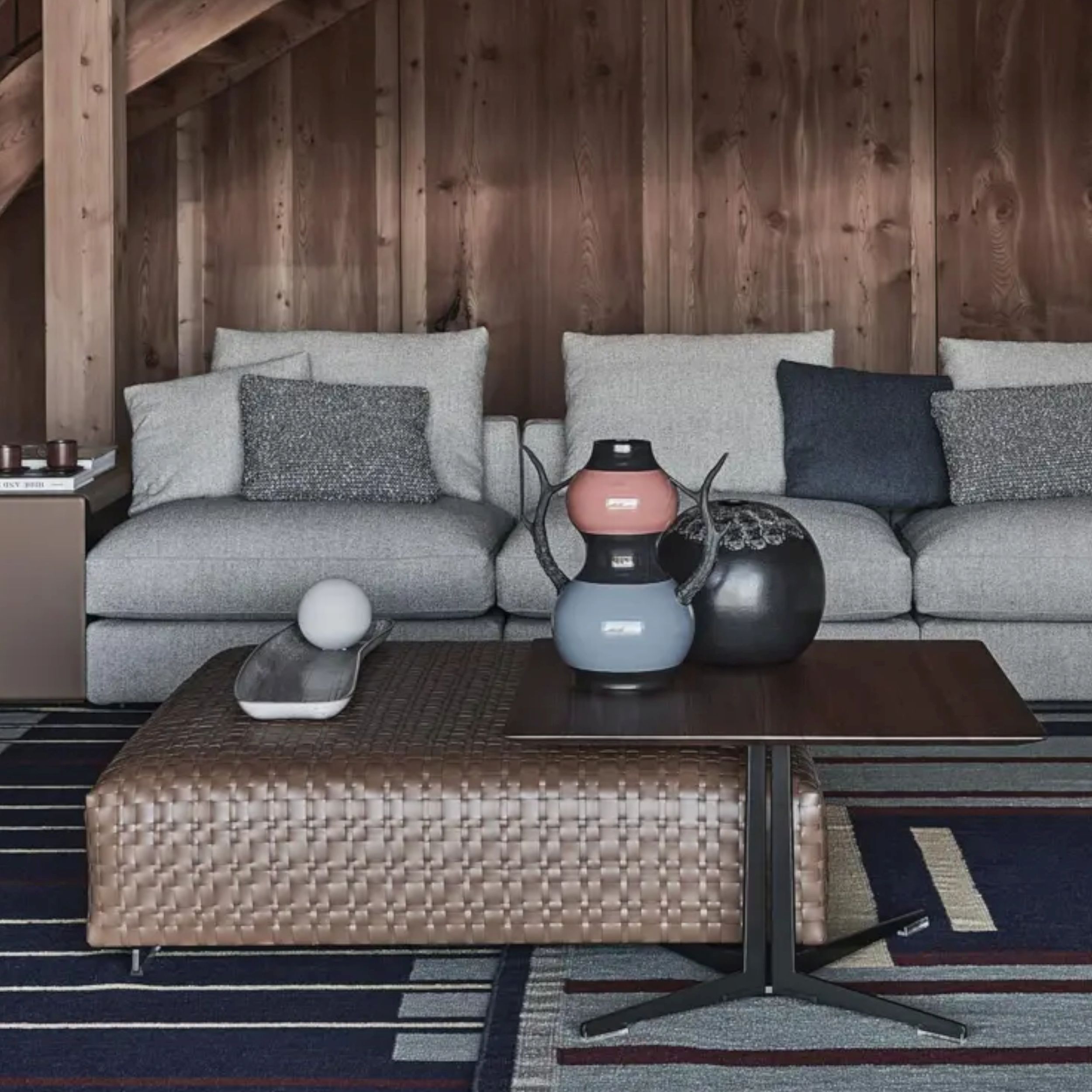 Ein gemütliches Wohnzimmer mit einem grauen Stoffsofa vor einer holzverkleideten Wand, ergänzt durch einen großen, geflochtenen Lederhocker in braun und einen Couchtisch, dekoriert einer bunten Vase.