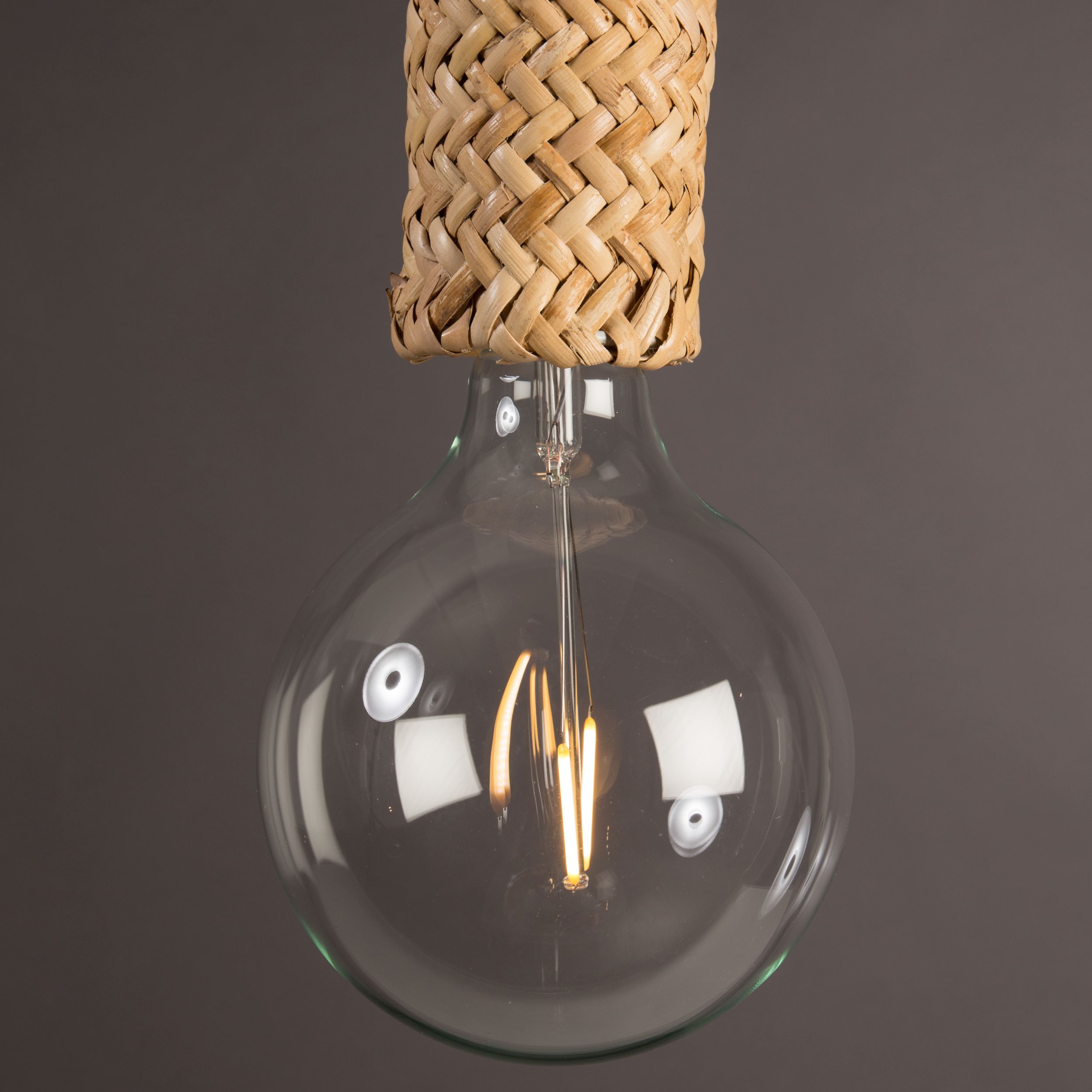 Nahaufnahme einer großen runden LED-Filament-Glühbirne an einer Hängelampe mit naturfarbenem Korbgeflecht vor dunkelgrauem Hintergrund.