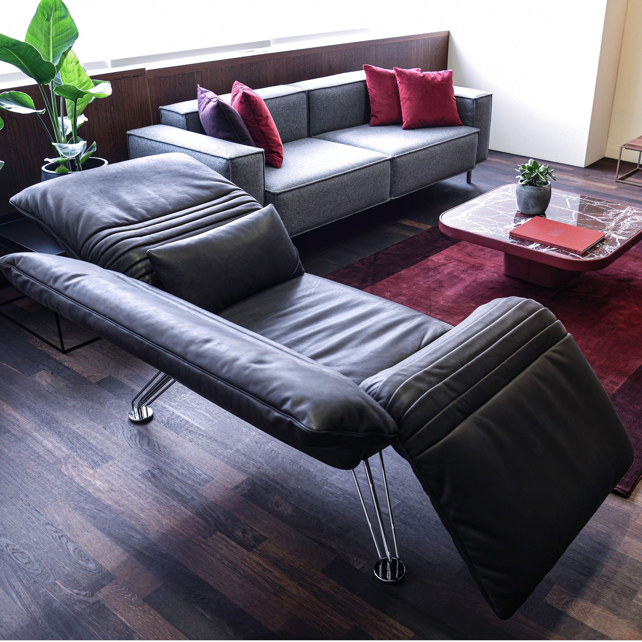 Ein modernes Wohnzimmer mit dunklem Parkettboden, ausgestattet mit einer schwarzledernen Relaxliege und einem kubischen grauen Sofa, ergänzt durch Kissen, Couchtisch und einen Teppich in rot.