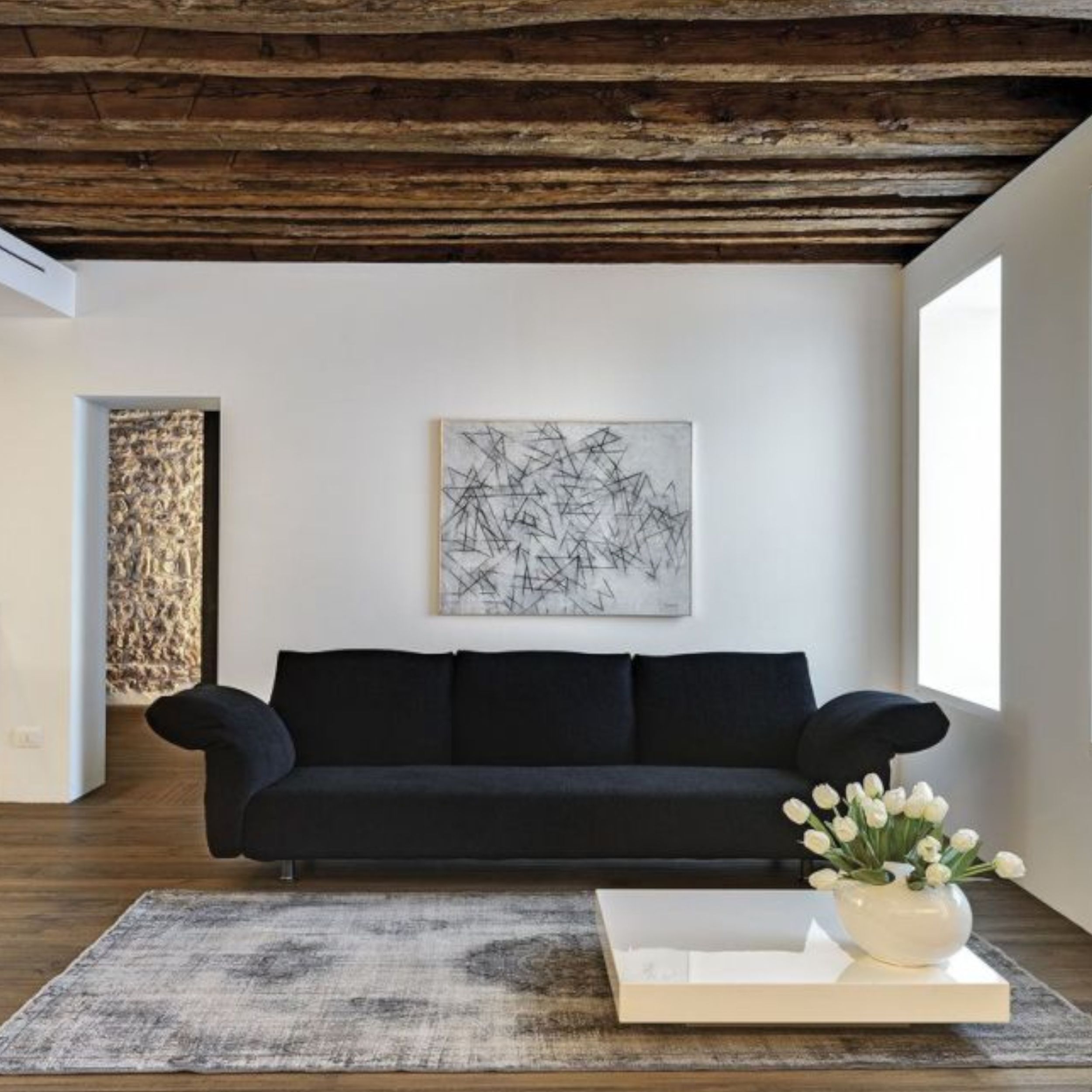 Ein gemütliches dunkles Stoffsofa steht in einem minimalistisch gestalteten Raum mit rustikaler Holzbalkendecke.